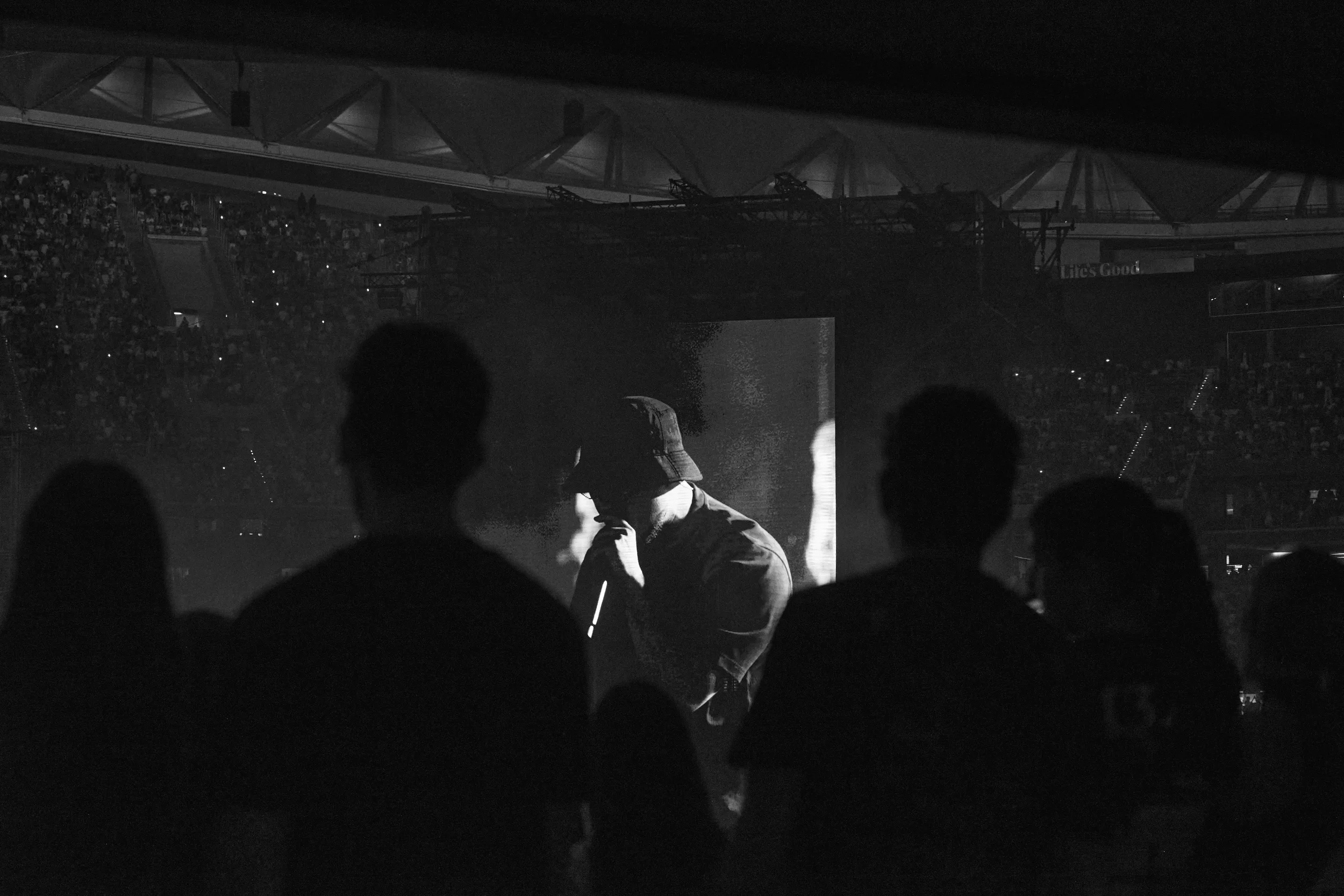 Personas en un evento oscuro con una pantalla grande y estructura de escenario visible, algunos espectadores en el fondo, en un ambiente de concierto o presentación.