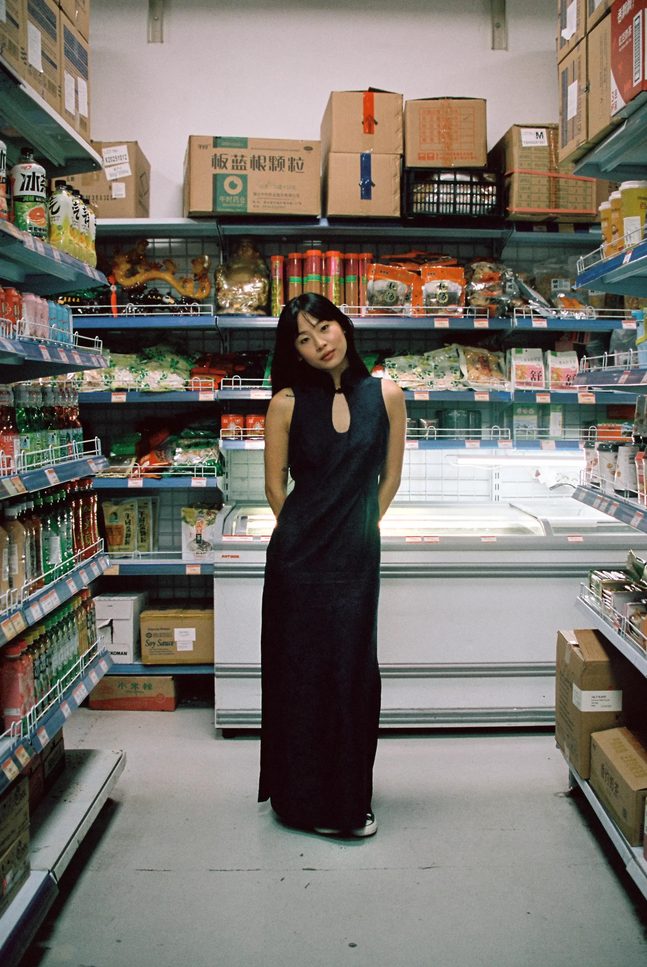 Una mujer joven con vestido largo y oscuro en una tienda de supermercado, mostrando un estilo elegante y moderno, rodeada de estantes con productos envasados y cajas en el fondo.