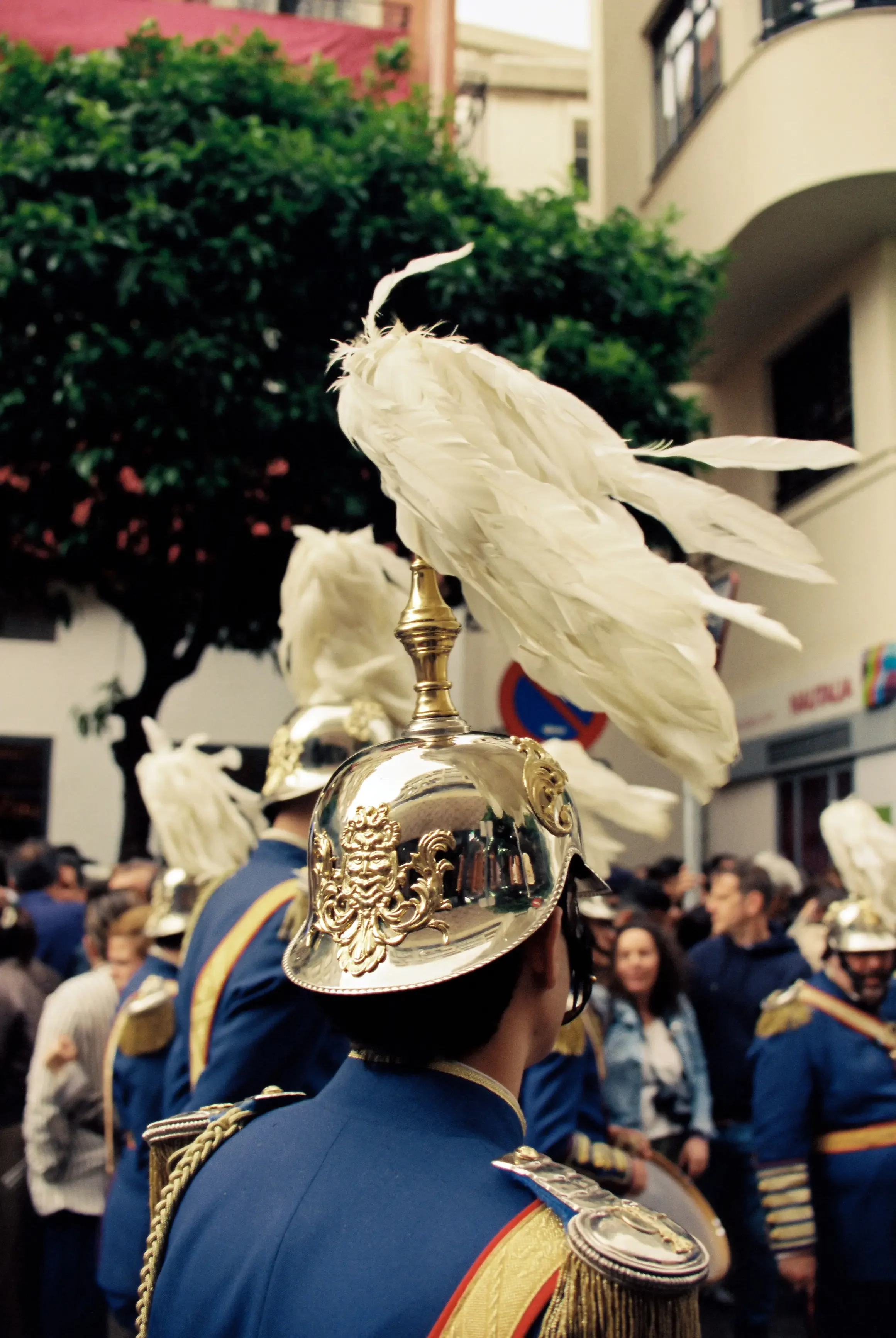 Uniformados con cascos decorados con plumas blancas y detalles dorados participan en una celebración en las calles de una ciudad, con edificios y árboles en el fondo, y muchas personas observando.
