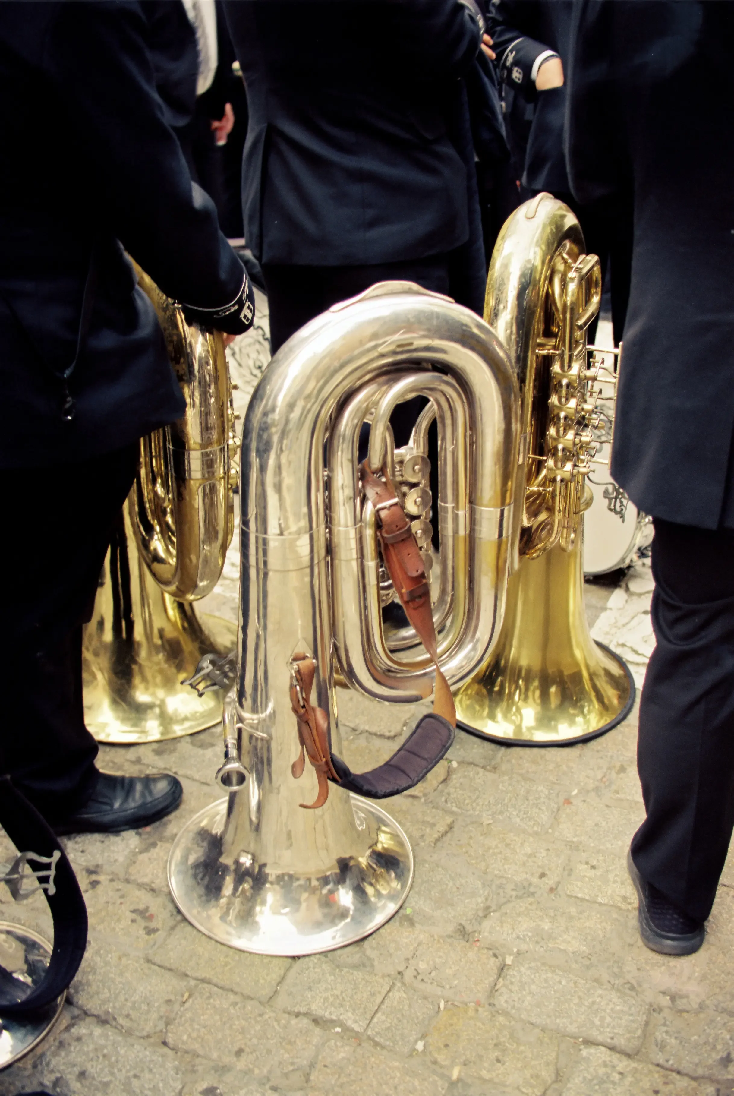 Grupo de personas con trajes oscuros y botas, rodeando y sosteniendo instrumentos musicales de bronce, como tubas y trombones, en una superficie de piedra.