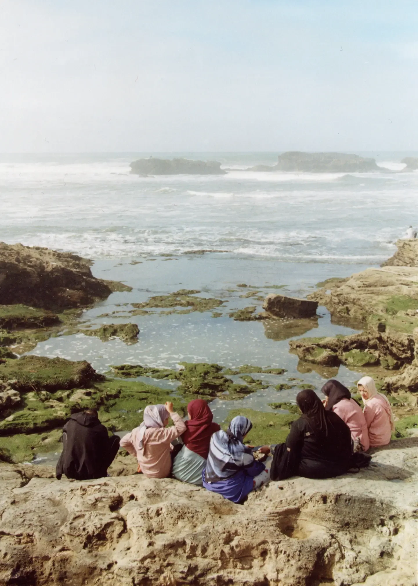 Grupo de personas sentadas en la playa, con rocas y mar en el fondo.