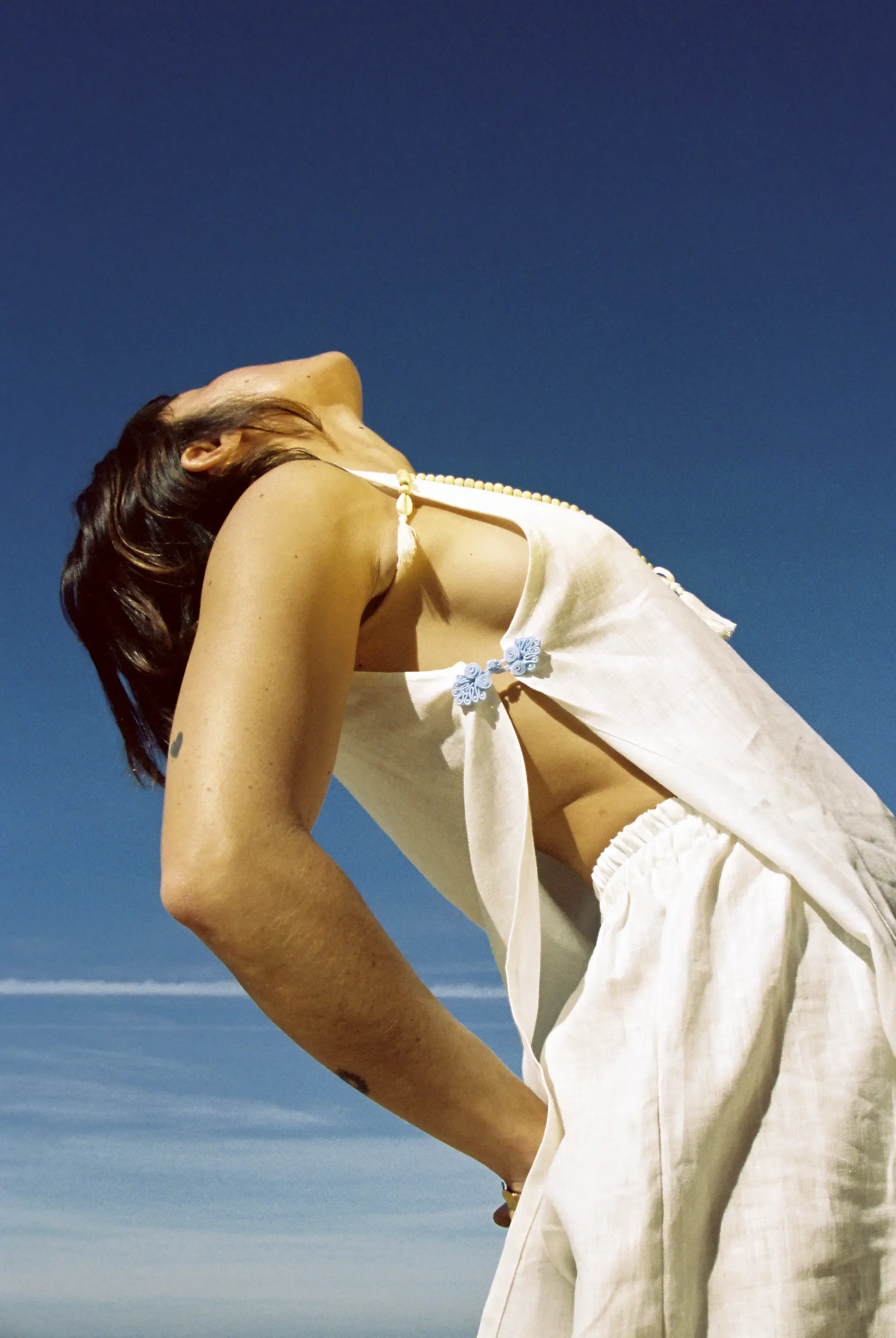 Mujer con blusa blanca y collar de perlas, de perfil, disfrutando del cielo azul despejado.