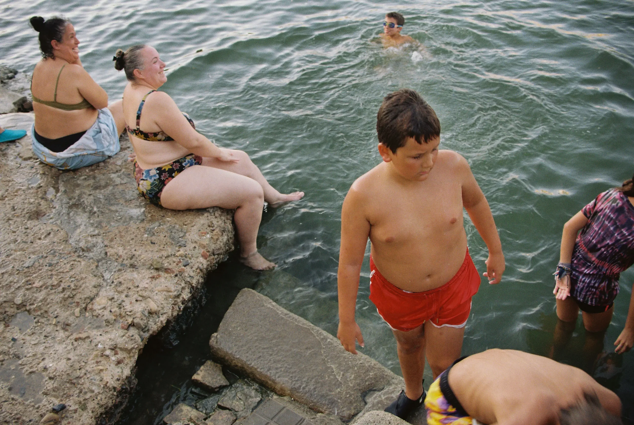 Personas adultas y niños en un muelle de piedra junto al agua, disfrutando del día en la playa, algunos bañándose y otros observando.