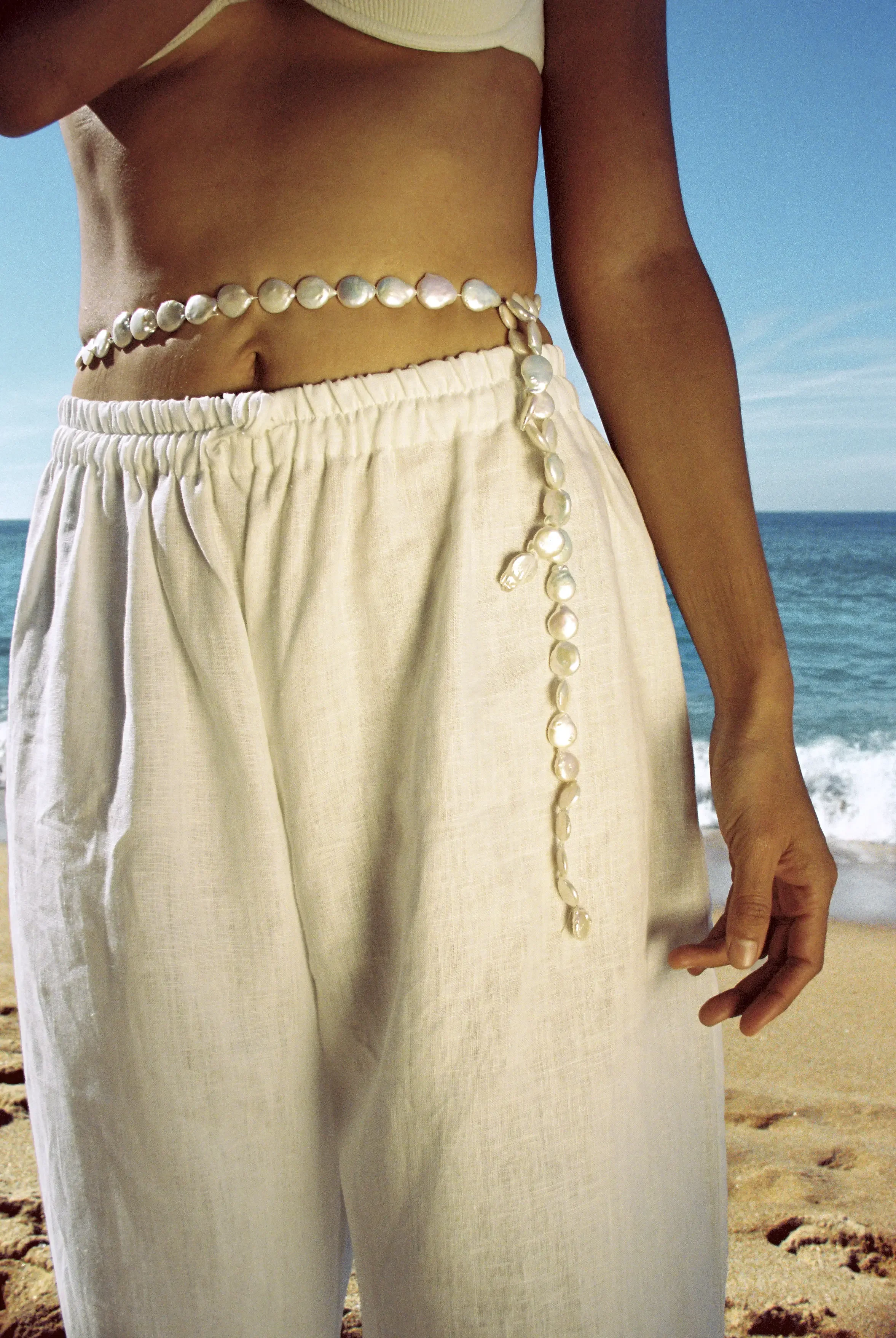 Persona con piel oscura usando un pantalón blanco de tela ligera en una playa, con el mar y el cielo azul de fondo.
