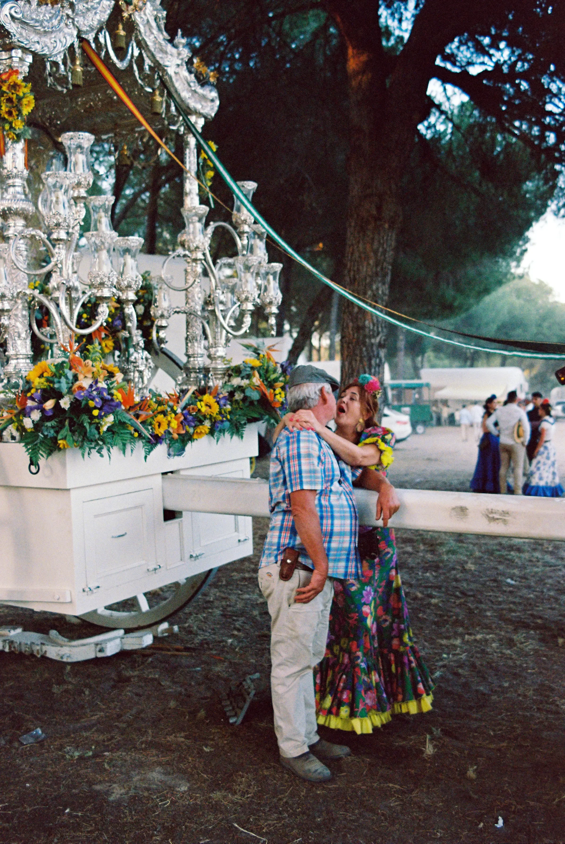 Pareja bailando en la feria, ella vestida con un vestido colorido y él con camisa de cuadros, junto a un carro decorado con flores y candelabros plateados.