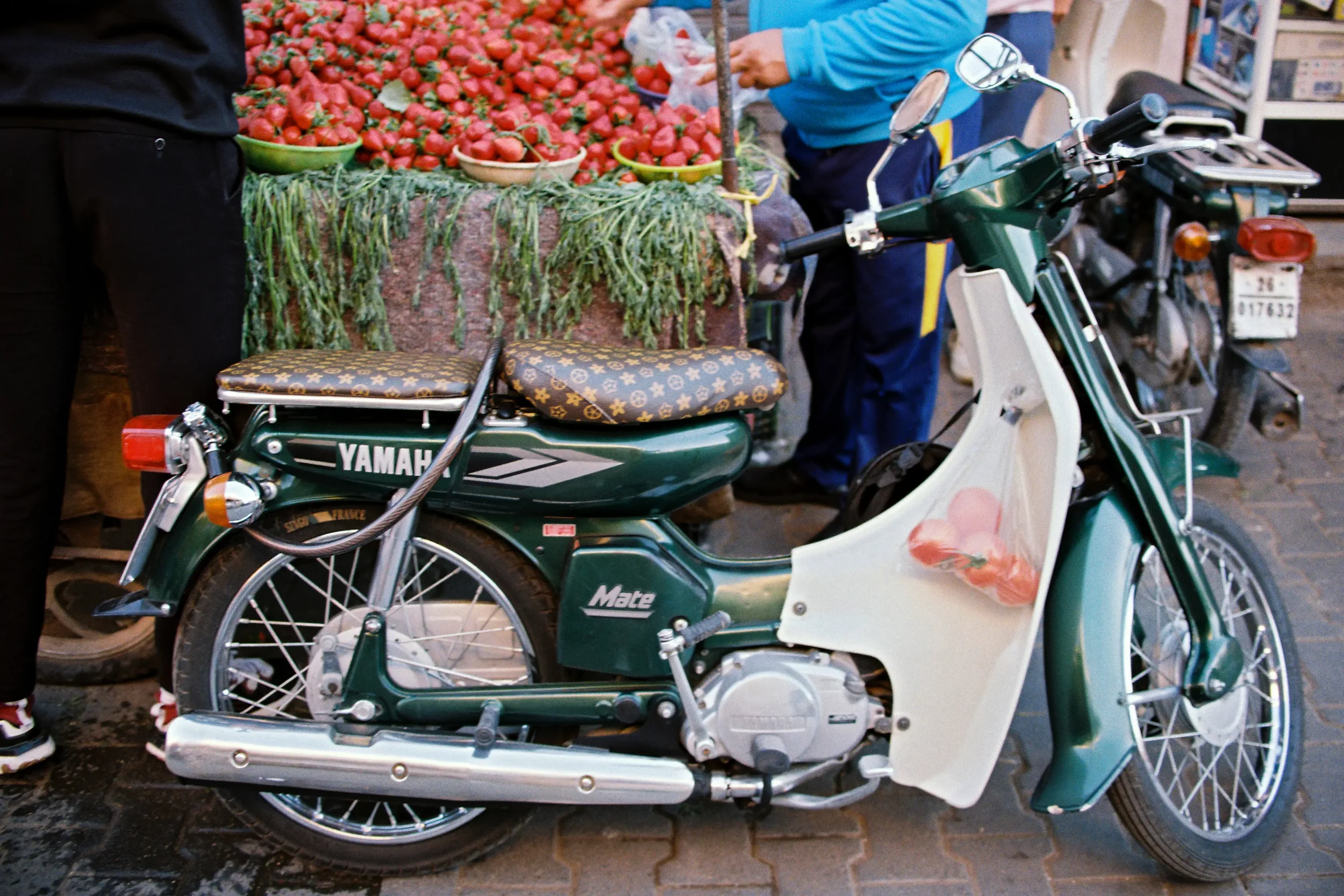 Motocicleta estacionada frente a puesto de venta de fresas en un mercado, con personas comprando.