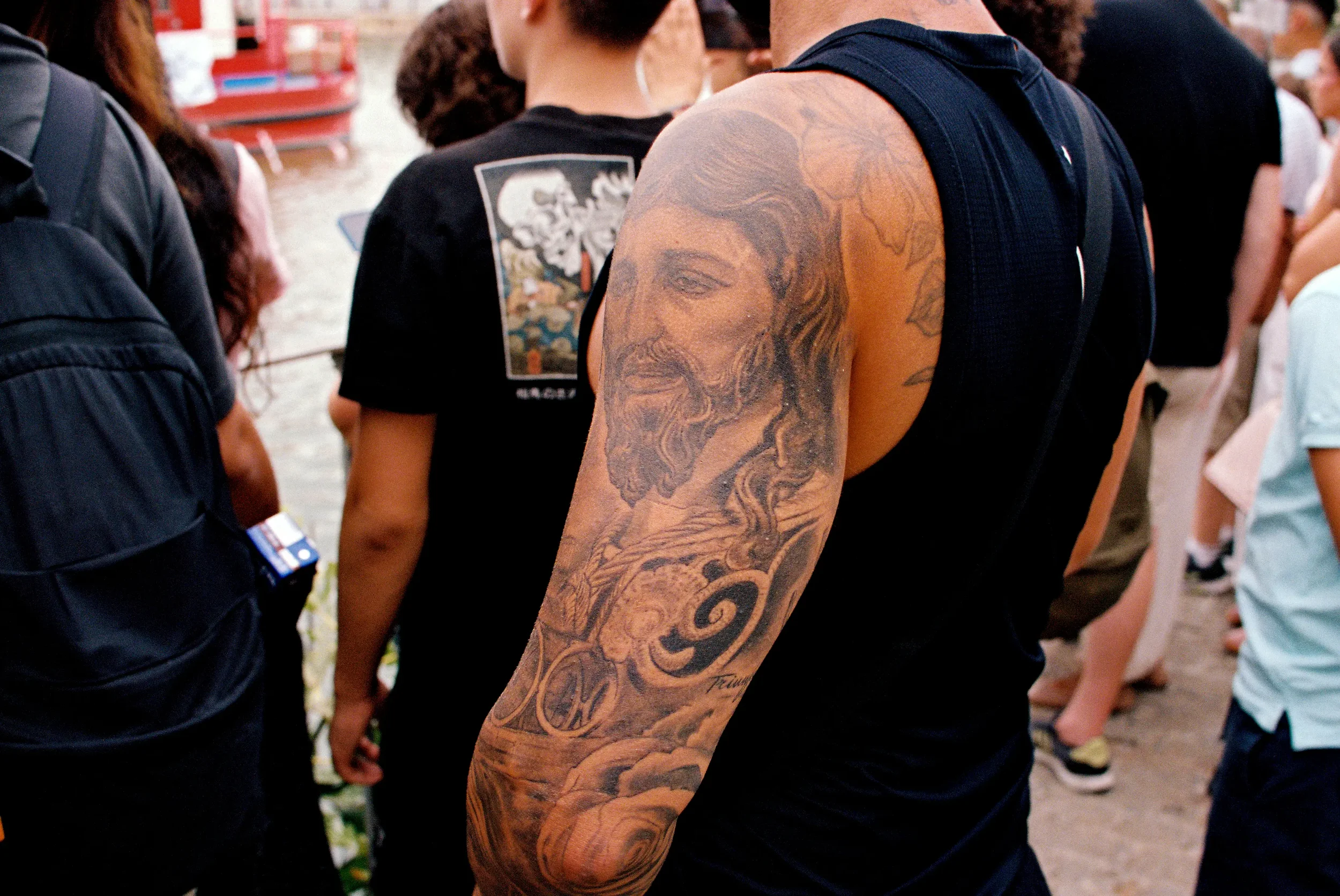 Brazos de varias personas en una multitud, una persona tiene un tatuaje grande de un rostro femenino en el brazo.