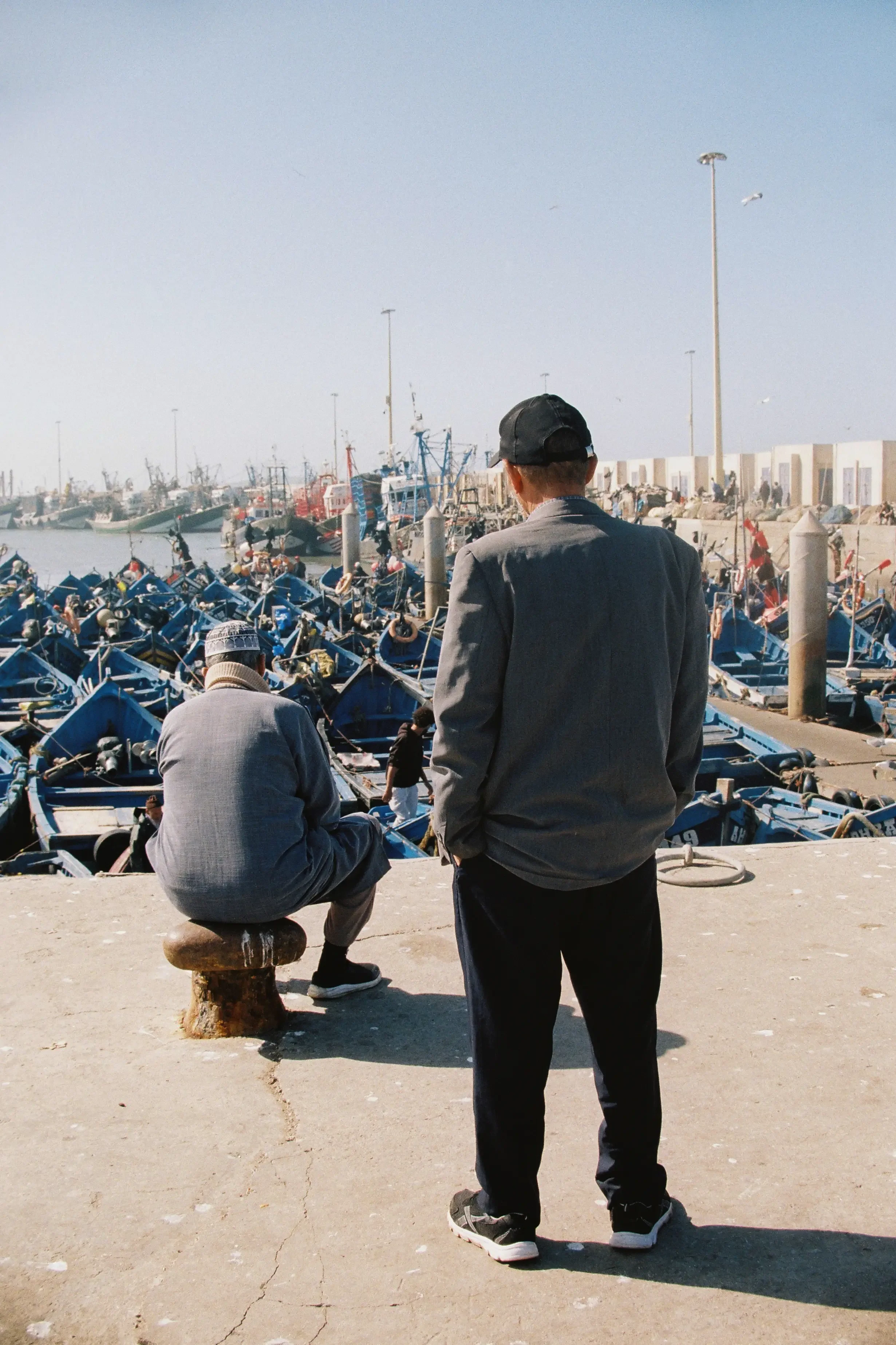 Dos hombres en un muelle observando botes y barcos en el agua, uno sentado en un banquito y el otro de pie con gorra, en una escena de día soleado.