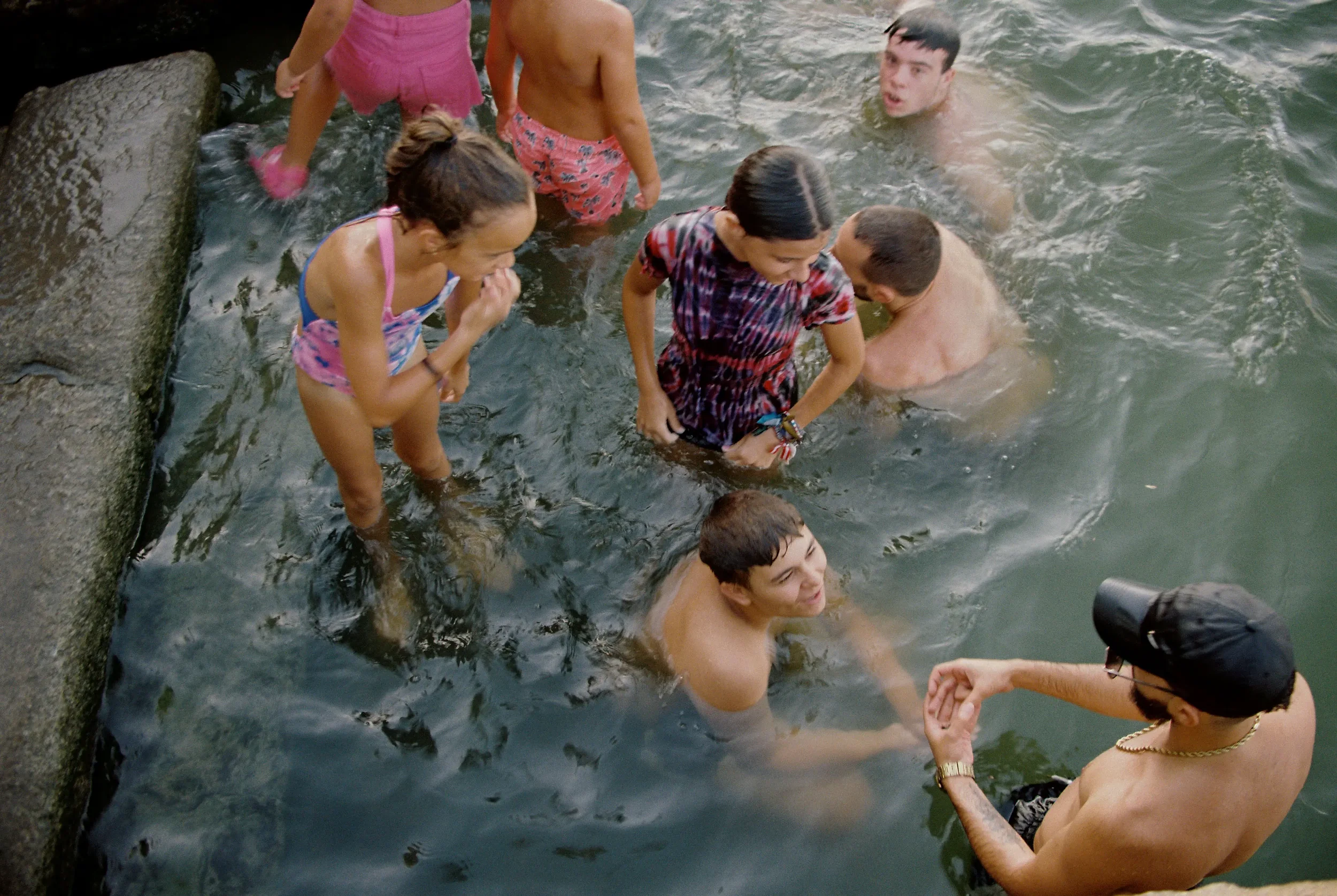 Grupo de niños y adolescentes en una zona de agua, algunos adultos ayudando y observando, disfrutando de un día al aire libre en un entorno natural.