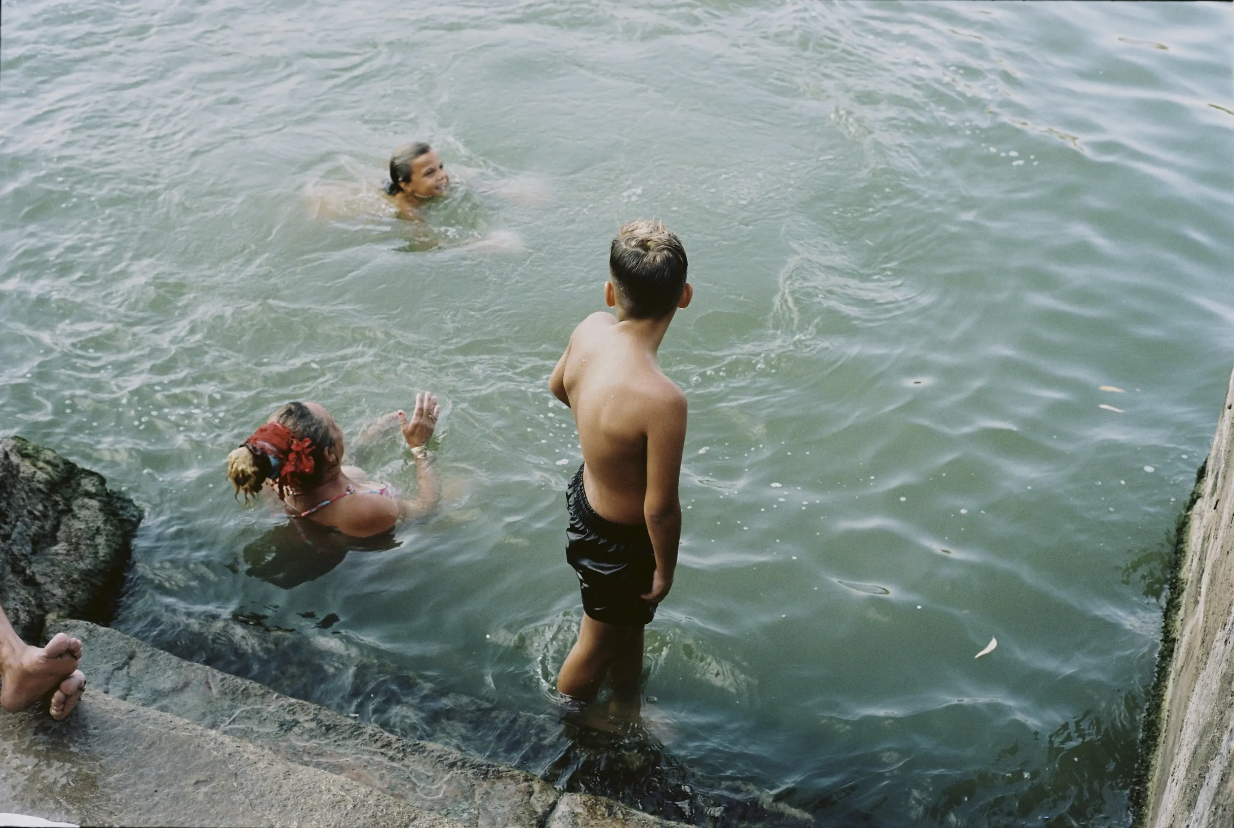 Niña y un joven jugando en el agua en la orilla del río, rodeados de rocas.