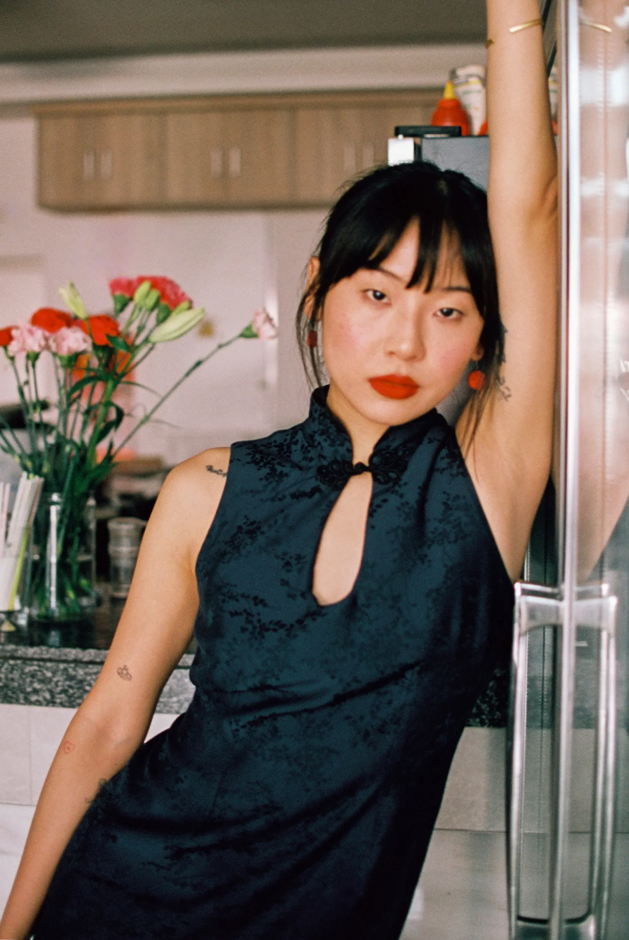 Joven mujer con cabello oscuro y corto, vestido negro sin mangas, con maquillaje rojo, en una cocina moderna, con flores en un florero