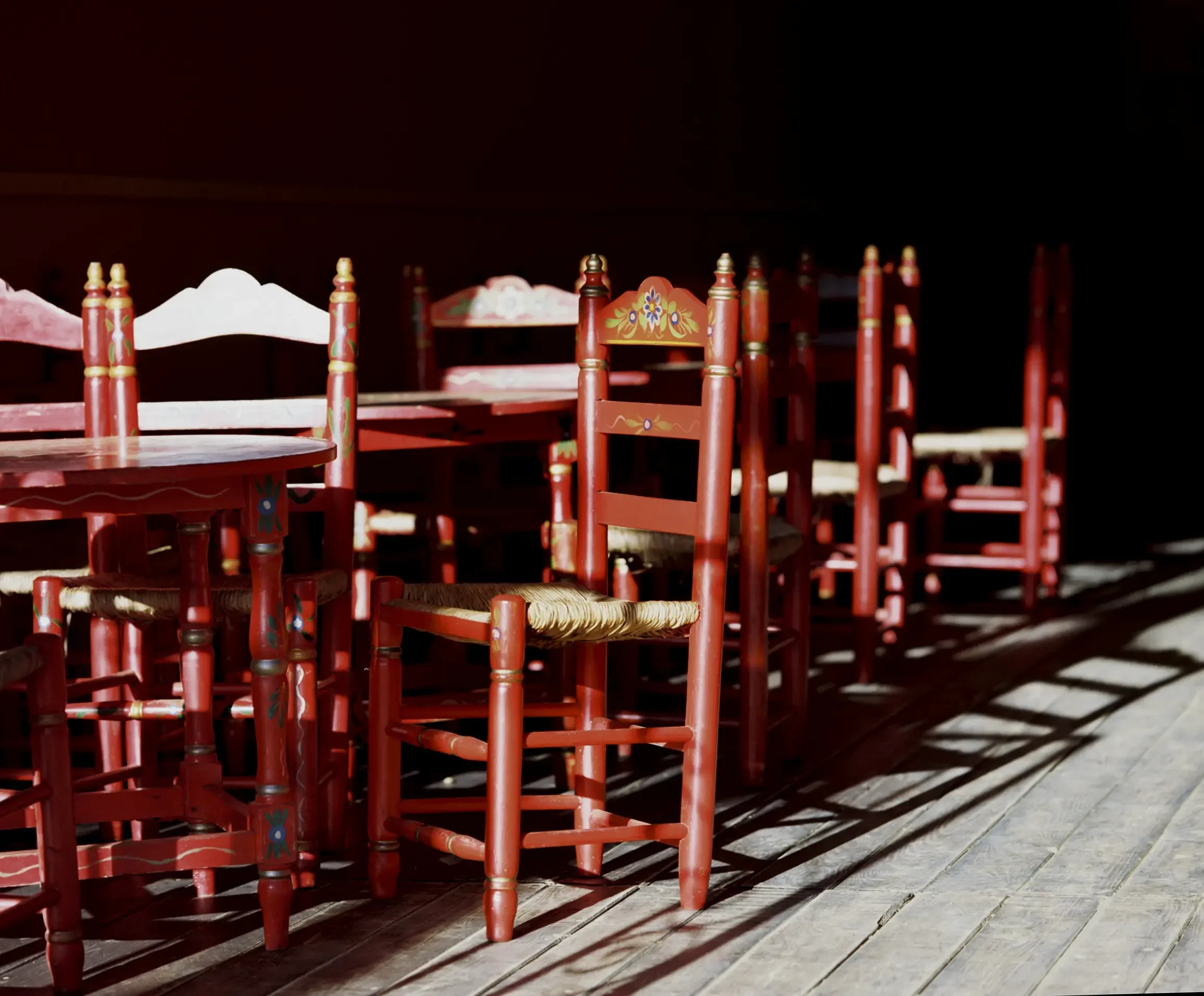 Filas de sillas de madera pintadas de rojo, decoradas con flores, en un espacio techado con largas sombras proyectadas en el suelo de madera.