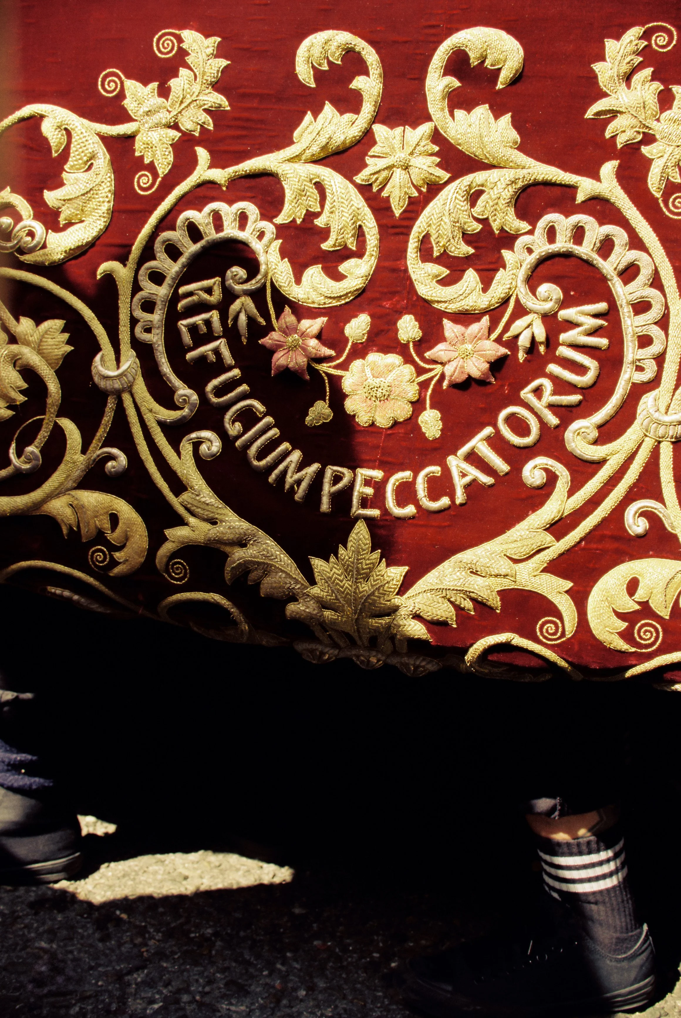 Brodería dorada y roja con la inscripción 'Refugio Impertinente', bordado con diseño floral y hojas decorativas, en la parte inferior las piernas de una persona con pantalones cortos y botas de senderismo.