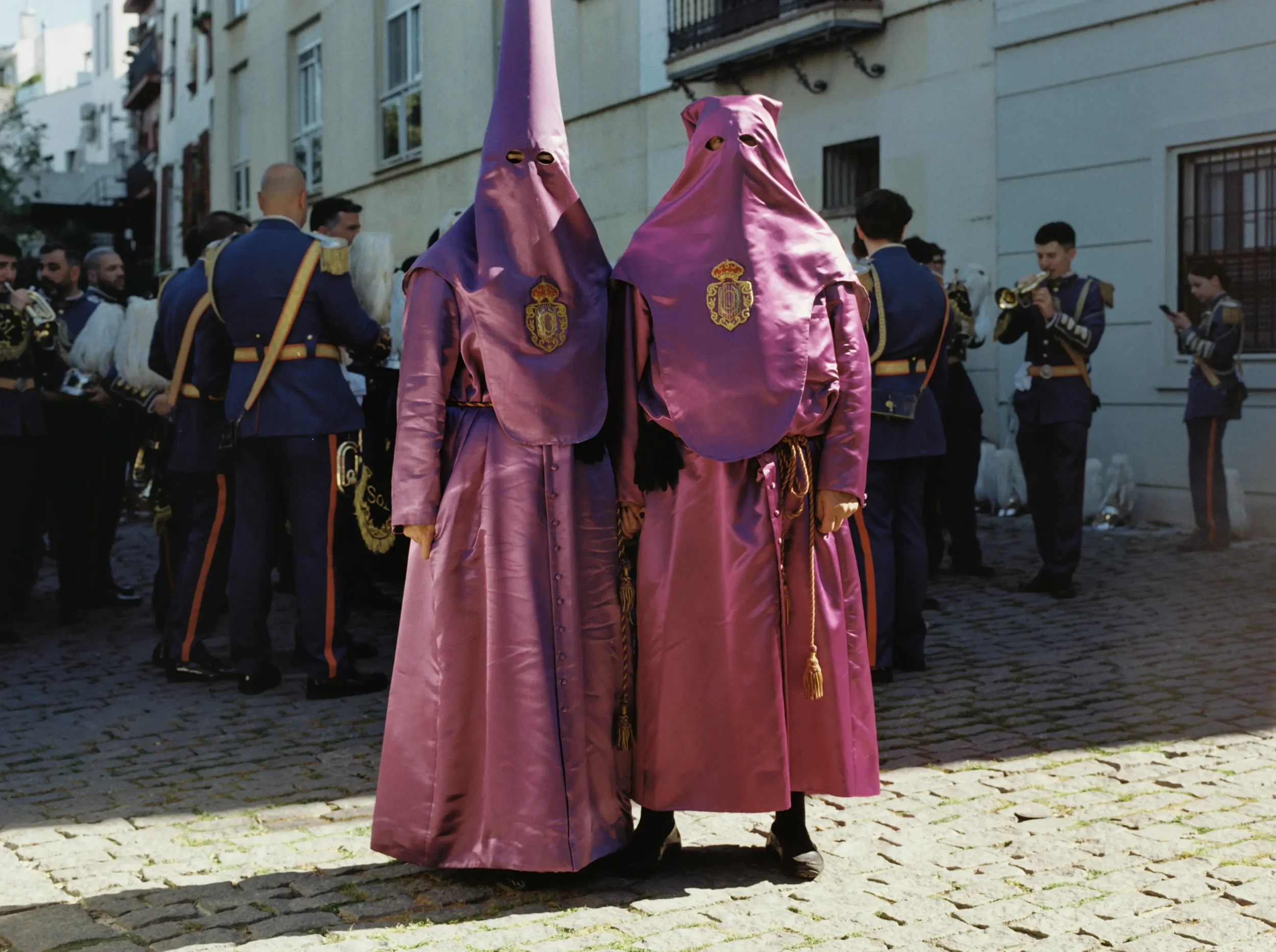 Dos personas vestidas con túnicas rosa y máscaras que parecen penachos en una protesta o desfile cultural en una calle adoquinada, con personas uniformadas observando.