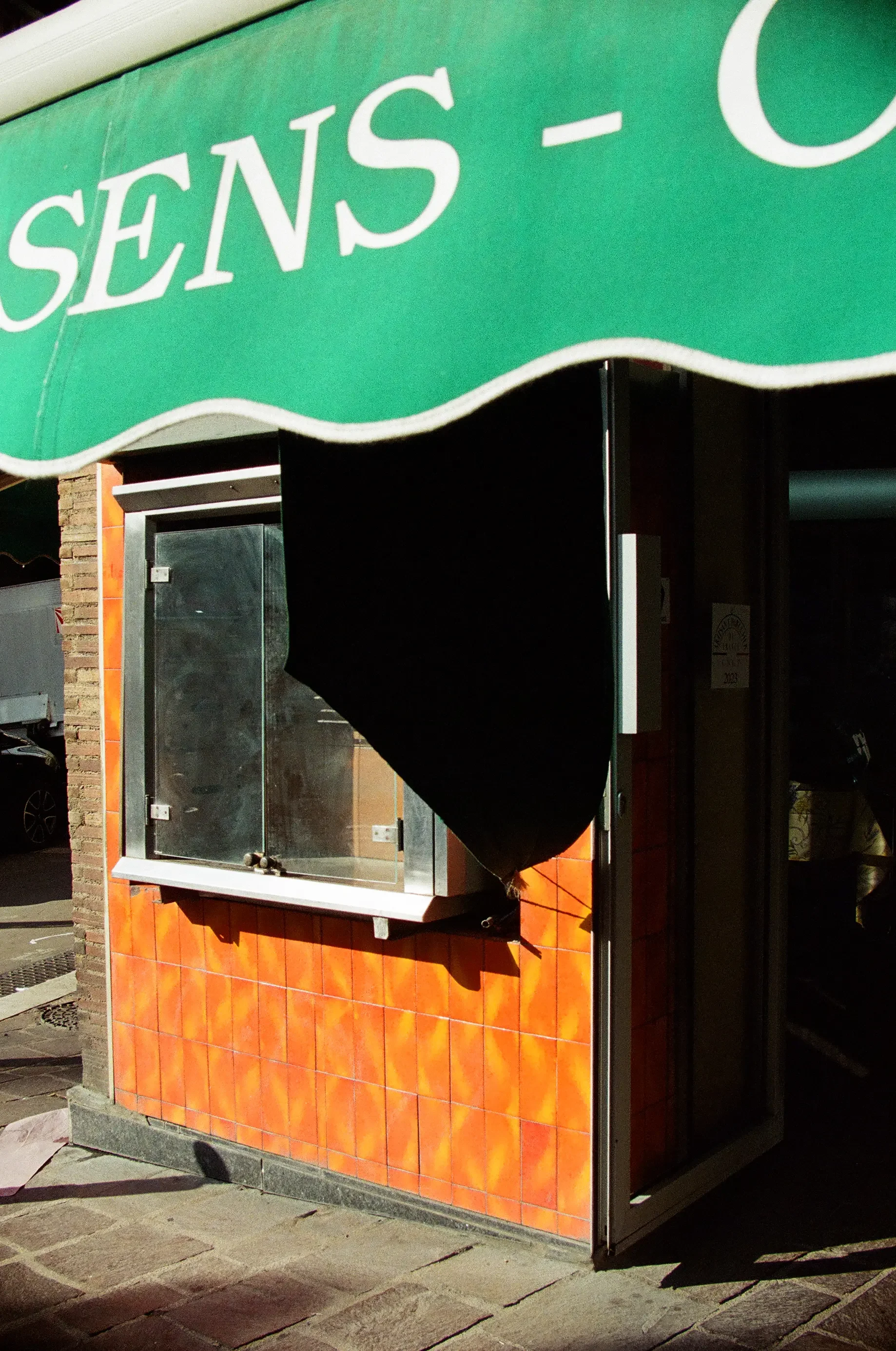 Vista de una fachada con un cartel verde que dice 'SENS-' y una ventana con protección de metal y madera en la parte inferior, en la ciudad.