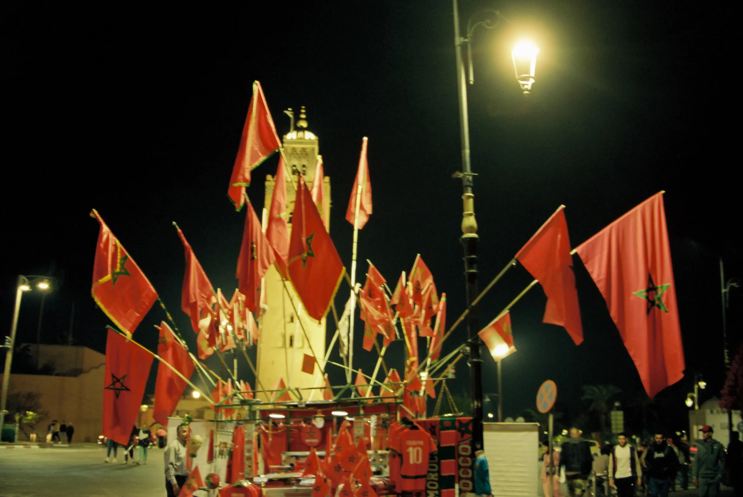 Banderas rojas con estrellas verdes ondeando en una plaza por la noche, con una torre y farolas iluminando el área.
