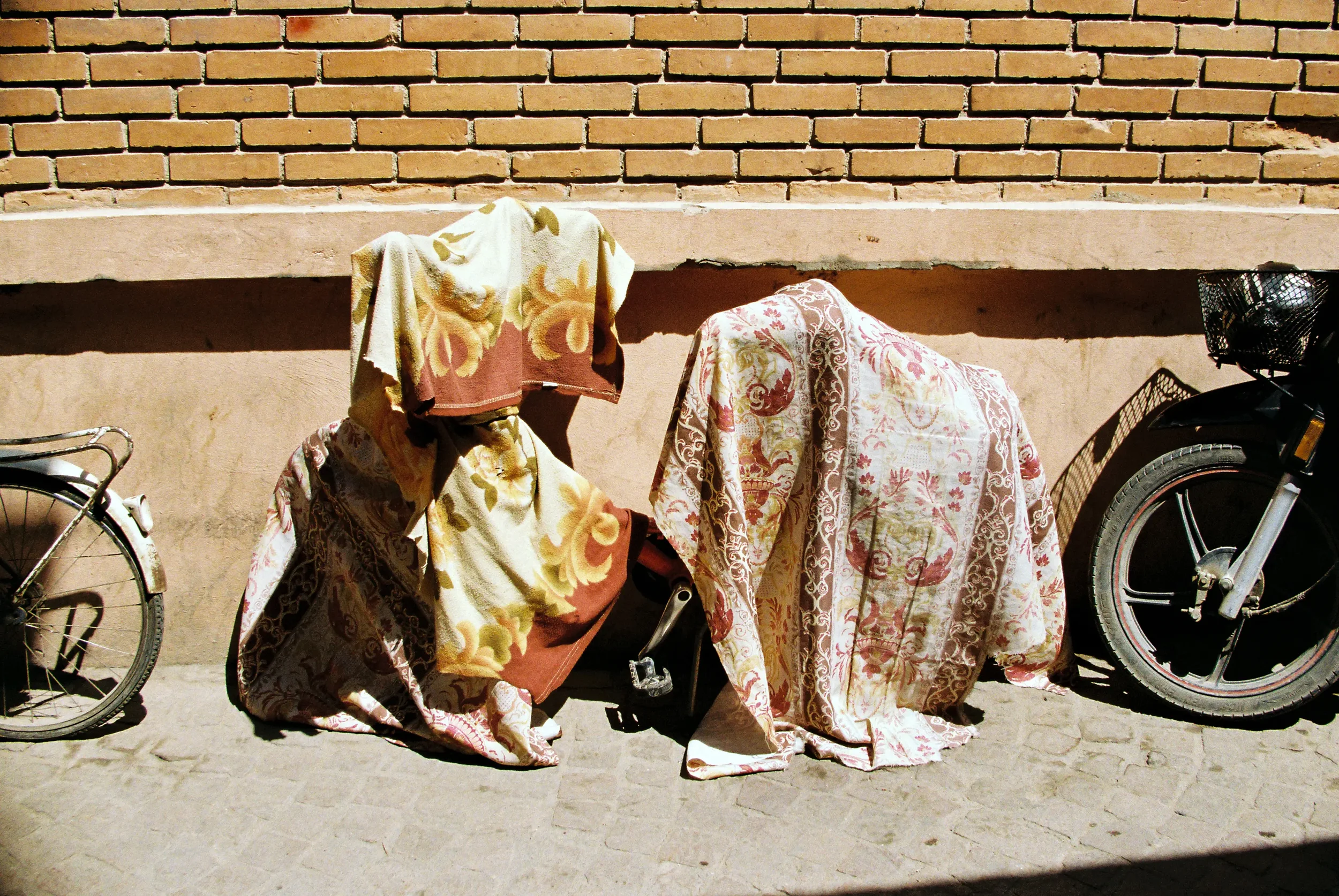 Dos bicicletas con cobijas cubriéndolas, apoyadas contra una pared con bloques de ladrillo y detalles en la base. La sombra proyecta los objetos. No hay personas visibles.