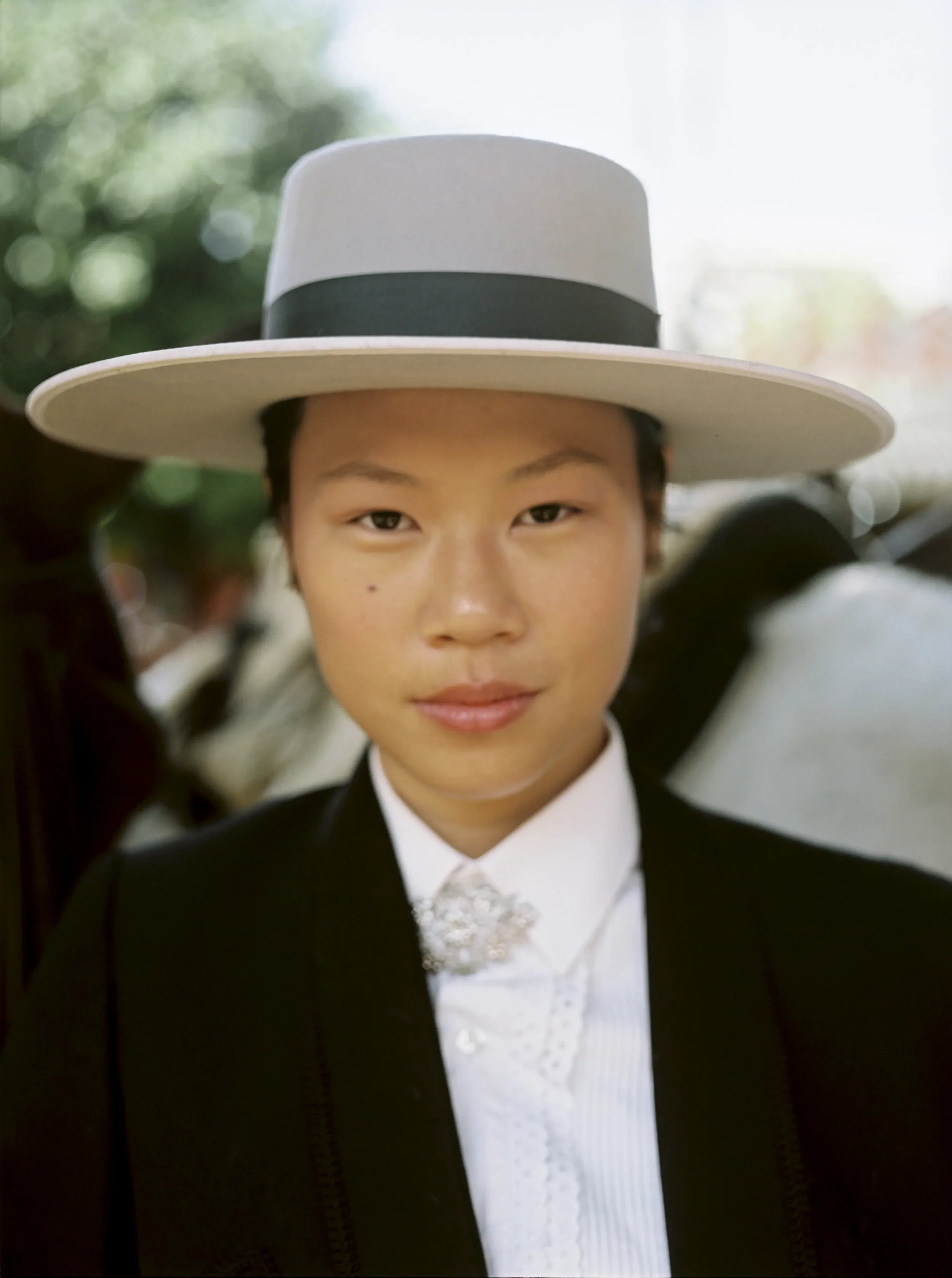 Primer plano de una persona con cabello corto, vestida con chaqueta negra, camisa blanca, y un moño de regalo en el cuello, usando un sombrero gris con banda negra, en un entorno exterior con árboles borrosos de fondo.