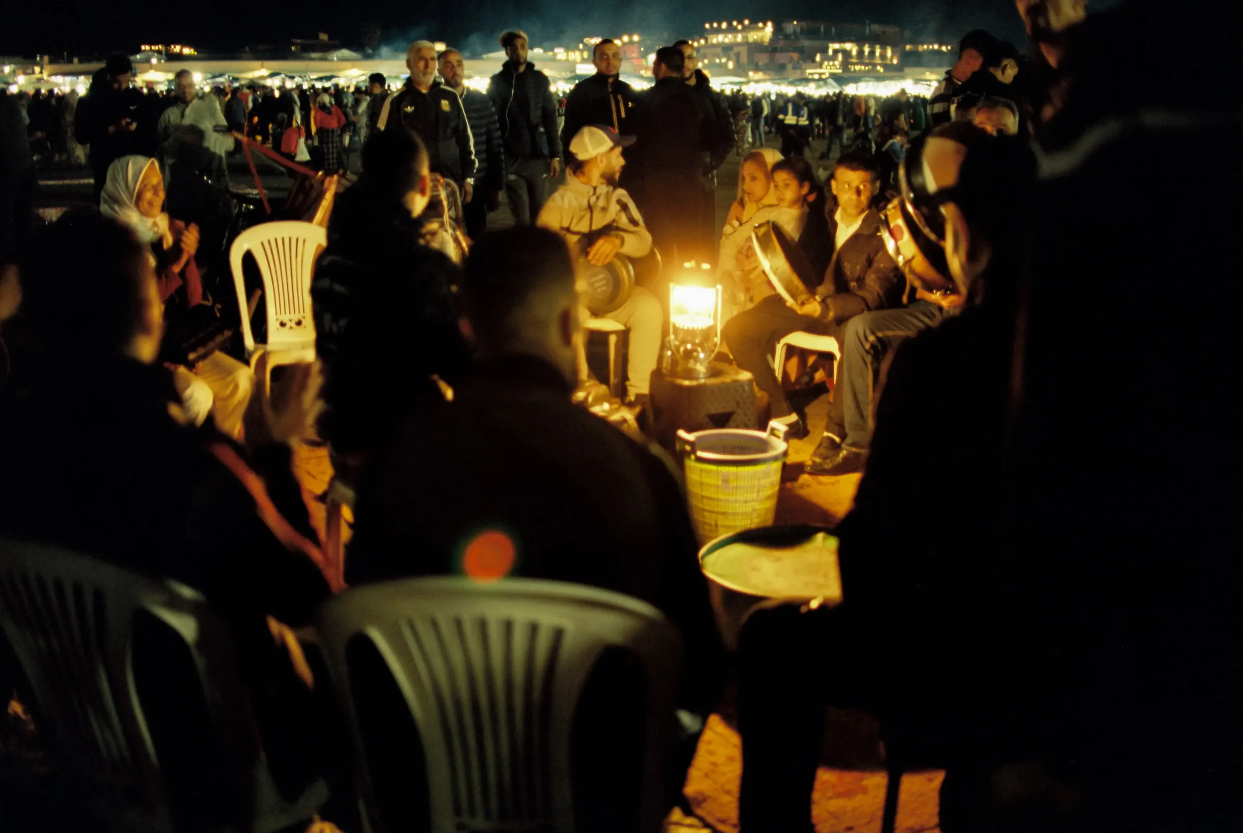 Personas reunidas alrededor de una lámpara en una noche en un evento al aire libre con muchas personas y puestos de comida y luces en el fondo.