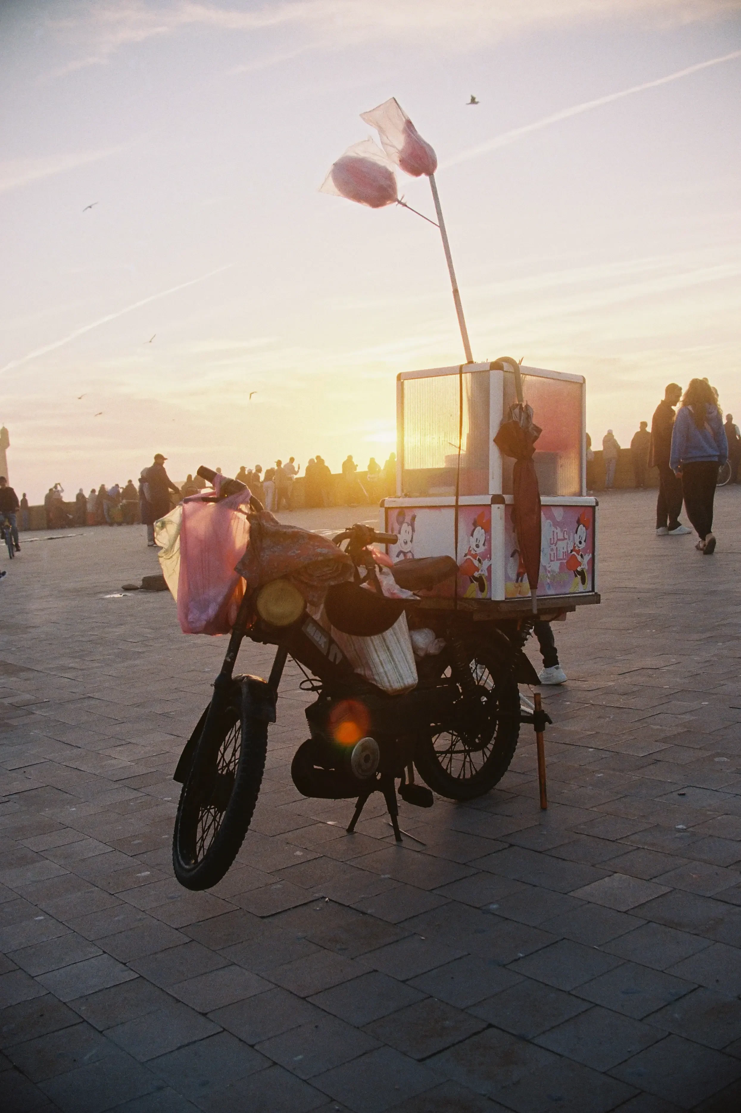 Una bicicleta con caja decorada con personajes de Mickey Mouse, cargada con bolsas y objetos, en un lugar público durante el atardecer, con personas caminando y un cielo con algunas nubes y aviones volando.