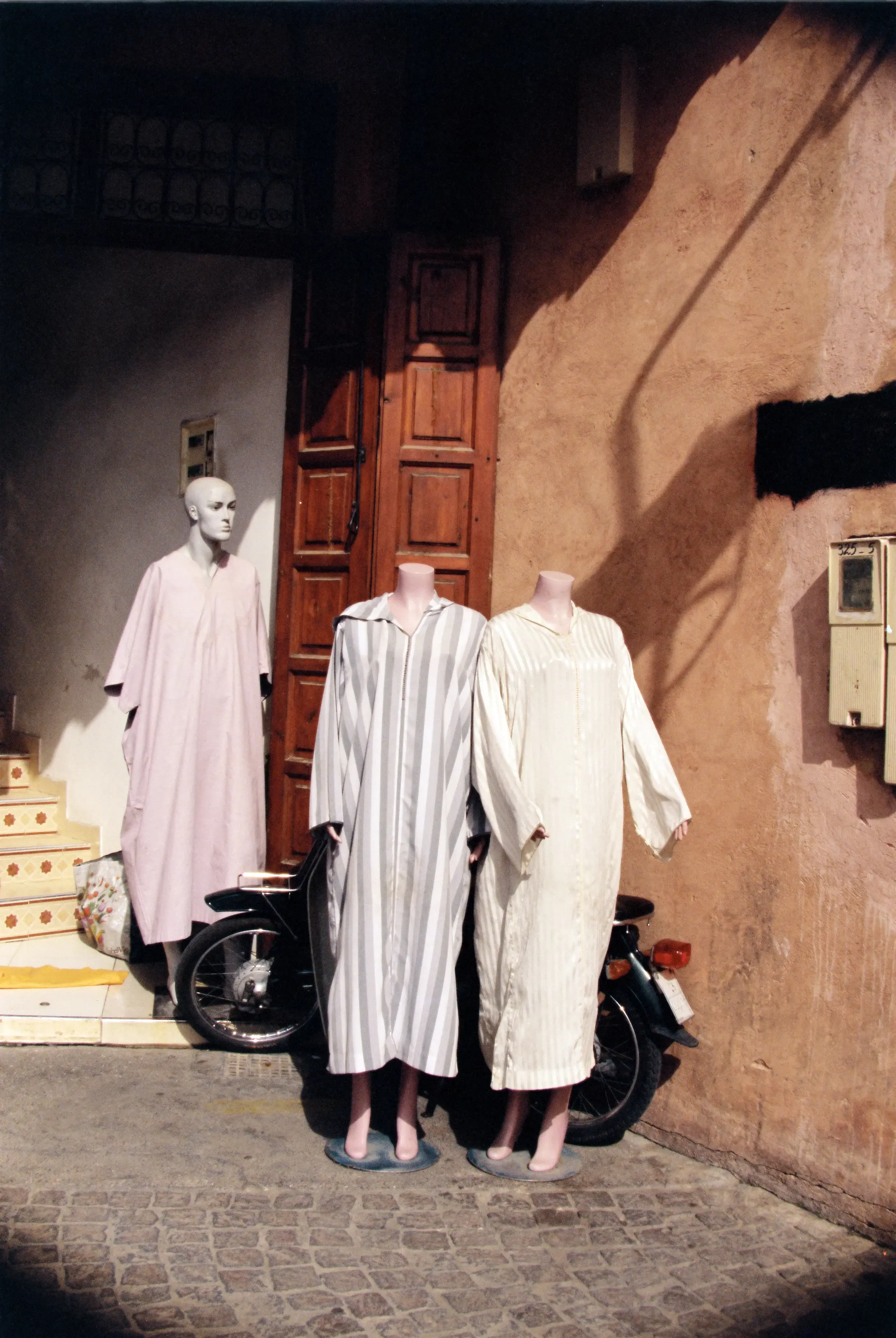 Cuatro maniquíes con pijamas de rayas blancas y grises en una acera frente a una pared de color tierra, con una motocicleta debajo de ellos, y una caja eléctrica en la pared.