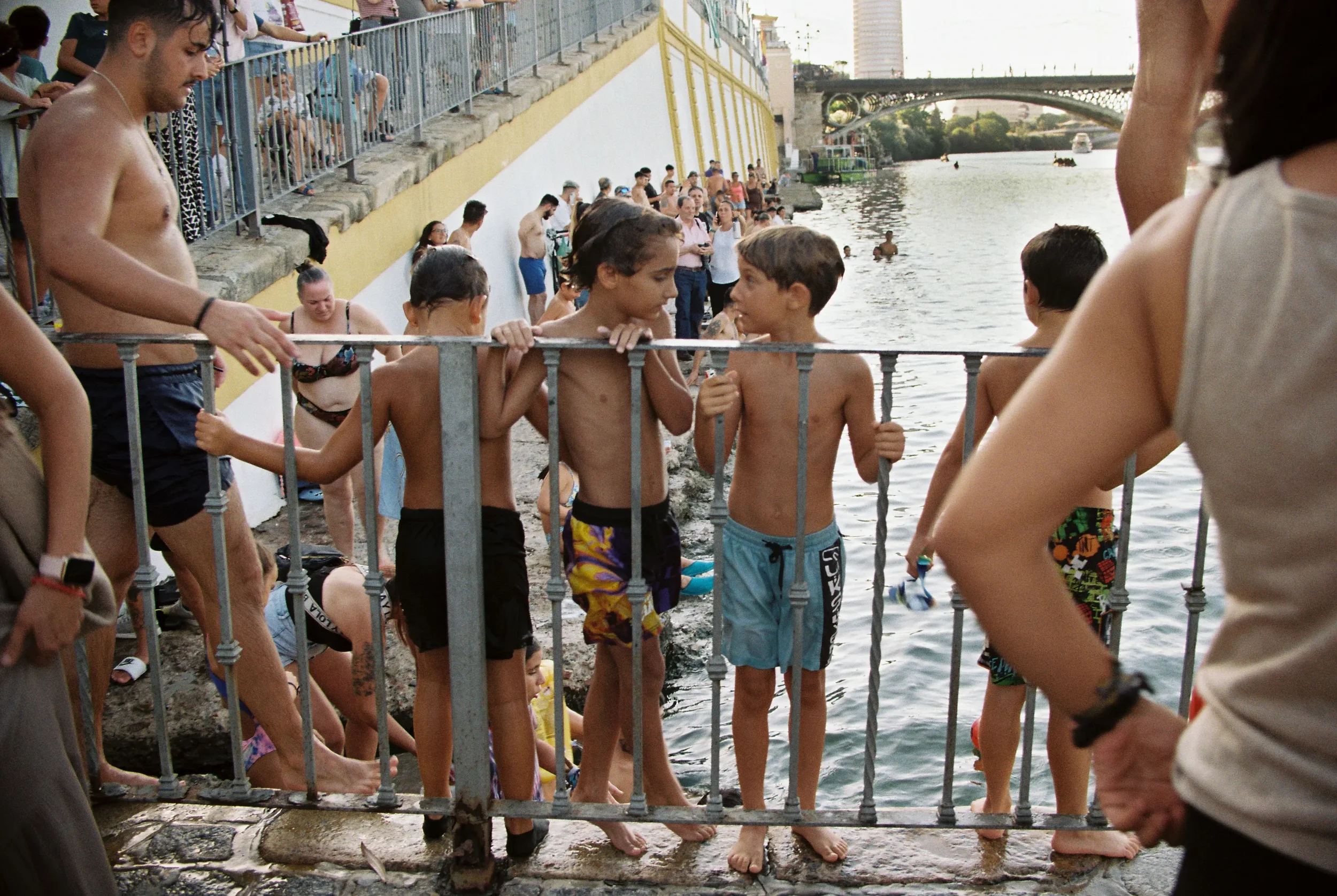 Muchos niños y adultos disfrutan bañándose en un río, algunos sin camiseta y otros con trajes de baño, mientras otros observan desde una barandilla de metal y una multitud de personas en el fondo cerca de una pared amarilla y un puente en la ciudad.