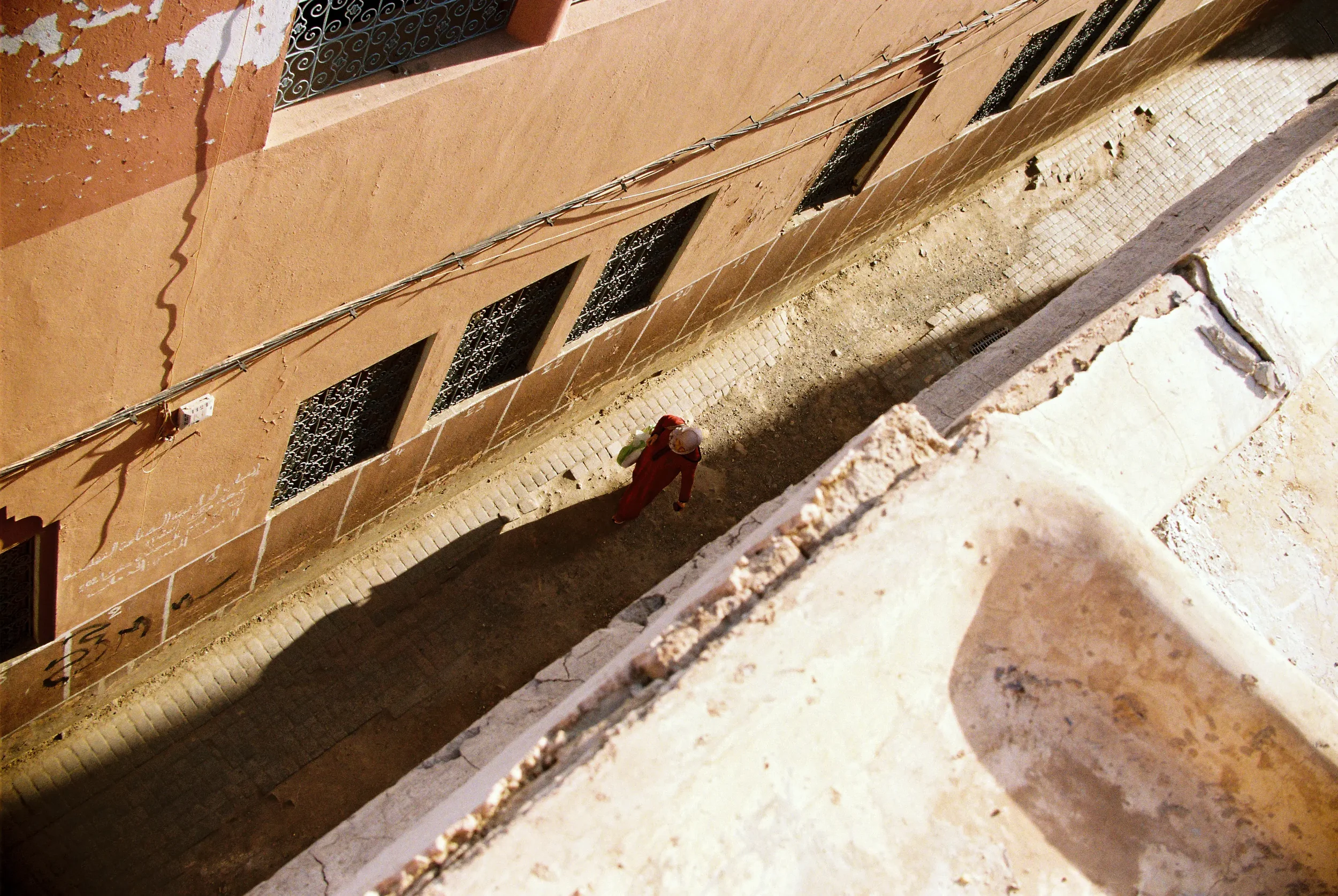 Vista desde arriba de un edificio con una pared de color terracota y ventanas con rejas negras. En la calle hay un trabajador con casco y uniforme rojo, y cables eléctricos paralelos a la pared.