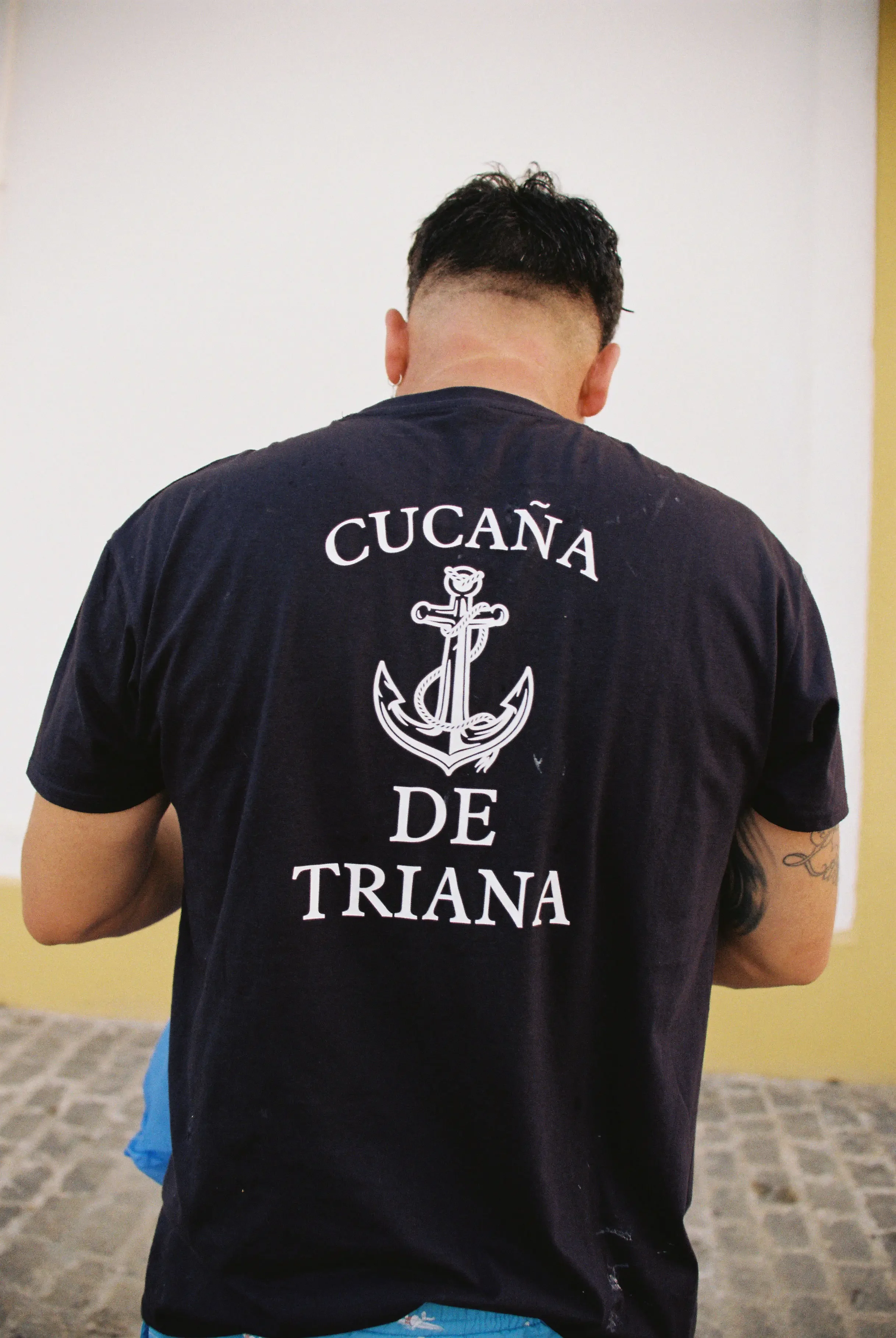 Persona vista de espaldas, con camiseta negra que tiene un ancla y letras en español, en un fondo de muro amarillo y piso de piedra.