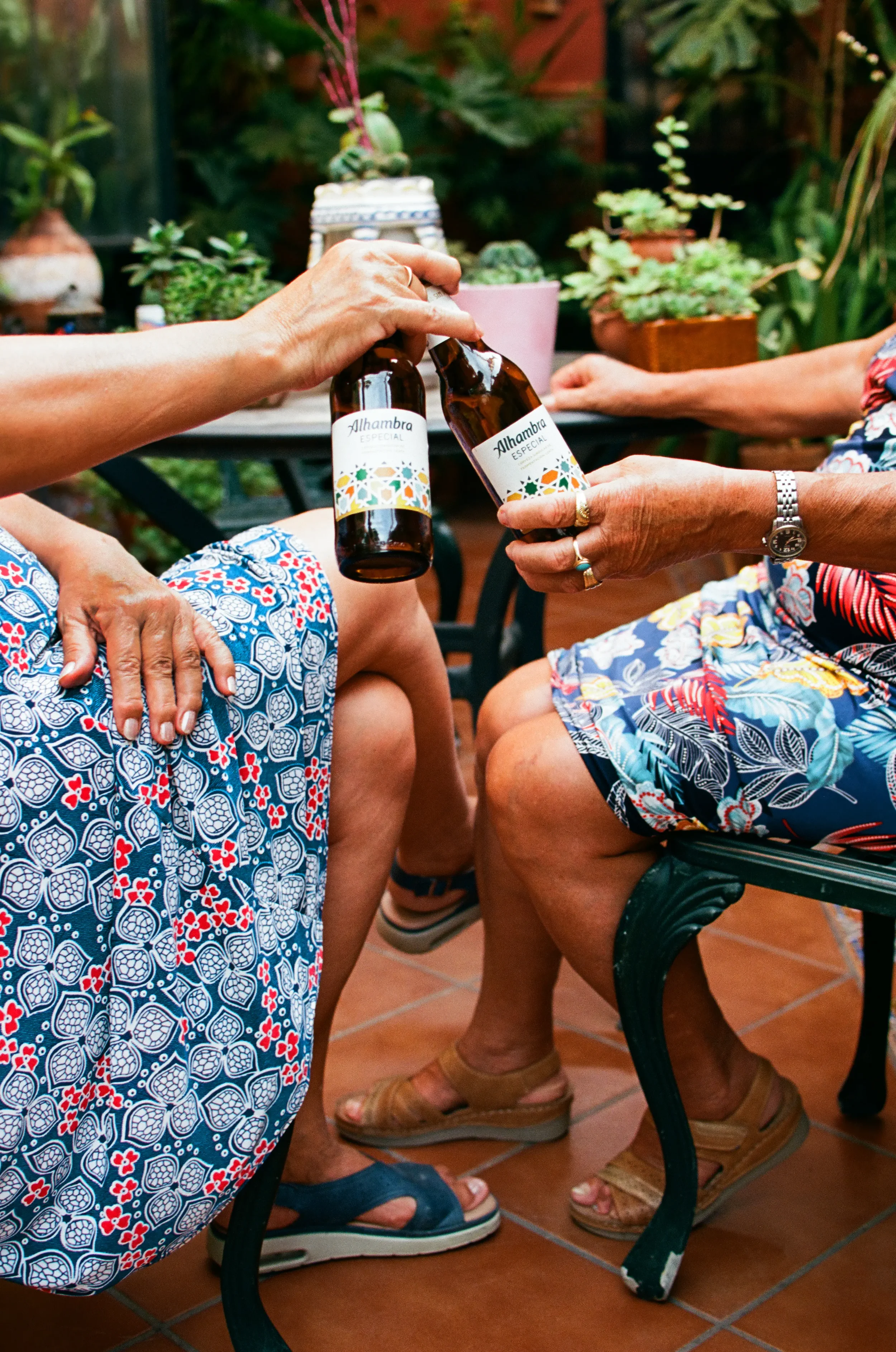 Personas intercambiando botellas de cerveza en una reunión social, con plantas en el fondo y personas sentadas con ropa colorida y sandalias en un espacio interior con piso de baldosas.