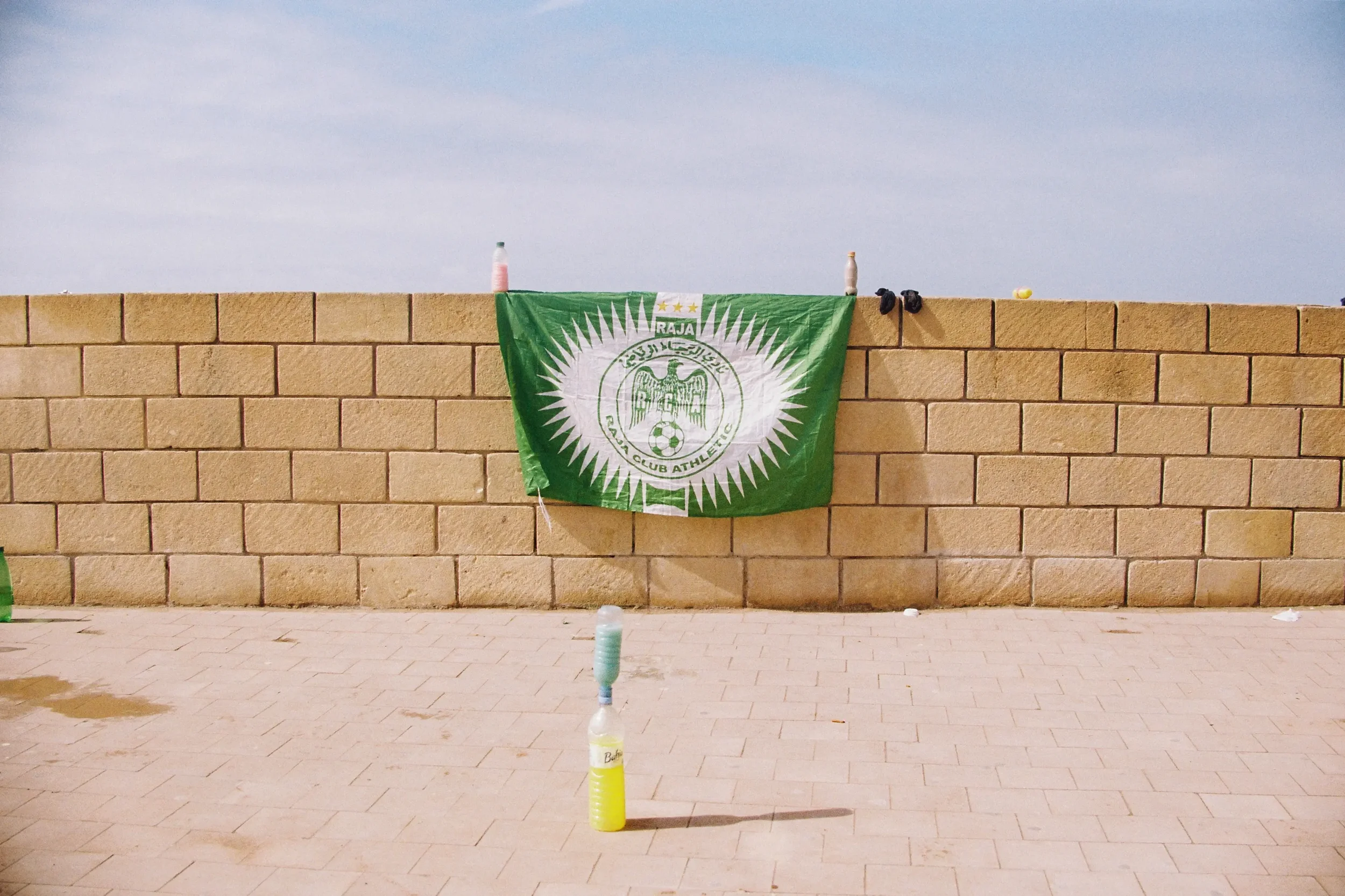 Una bandera de un club de fútbol deportivo colgada en una pared de bloques de piedra, con botellas de plástico encima de ella y en el suelo, y objetos dispersos en la parte superior. El fondo es un cielo despejado.