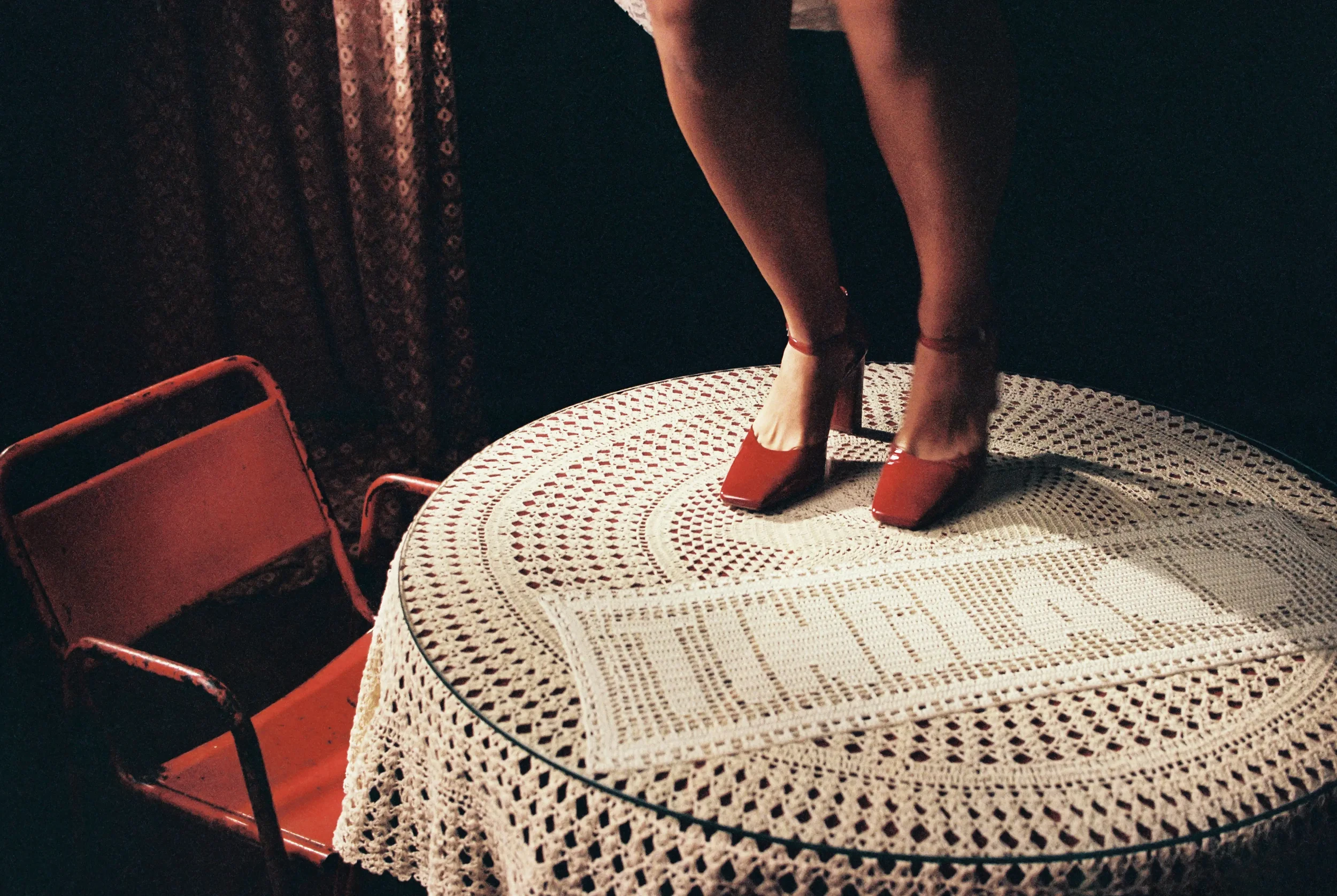 Esta imagen muestra las piernas de una persona con zapatos rojos de tacón alto, de pie sobre una mesa de mimbre blanca, en un entorno oscuro con cortinas con estampado floral y sillas rojas a su alrededor.