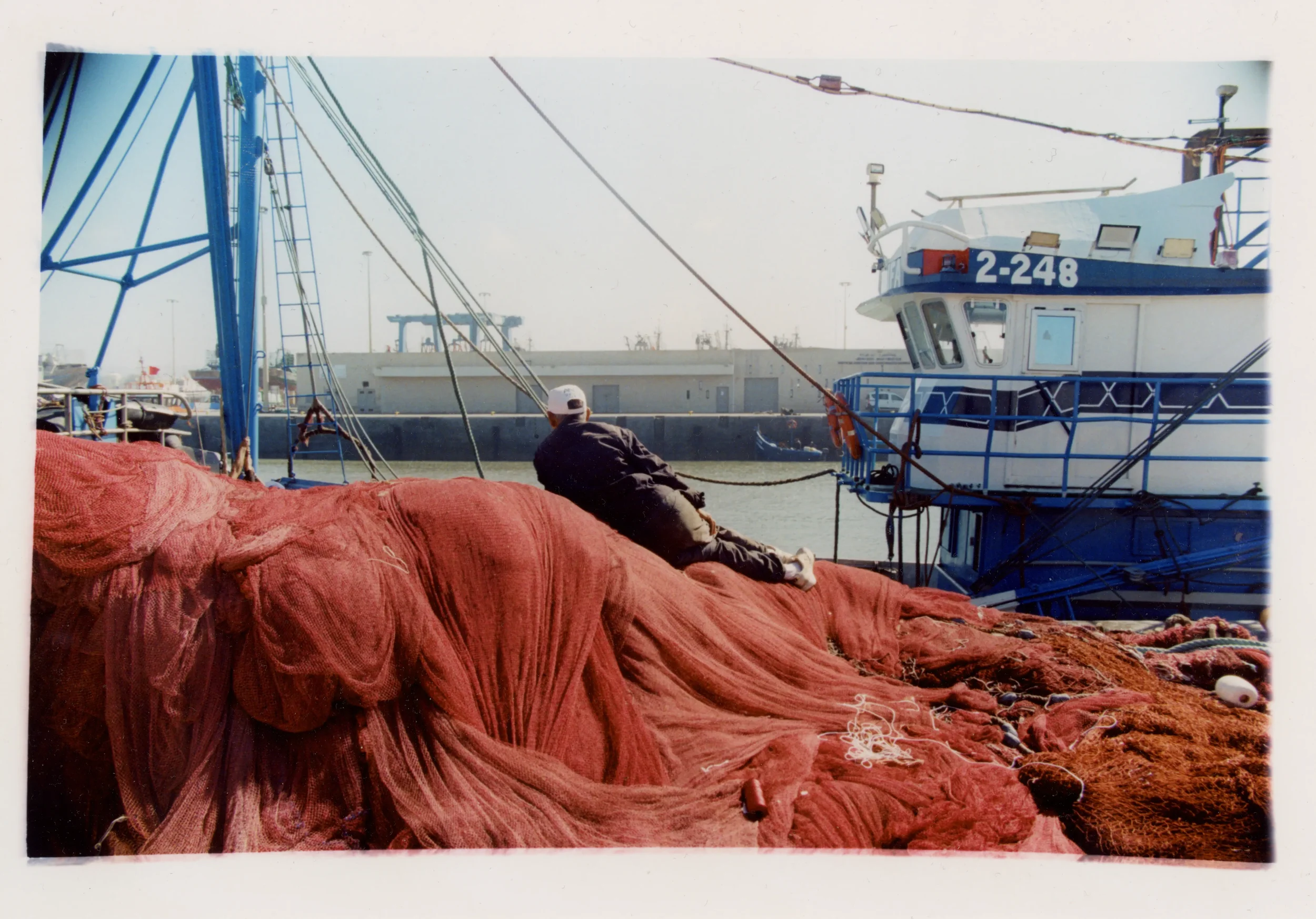 Persona descansando en un muelle con redes de pesca y un barco de fondo en un puerto.