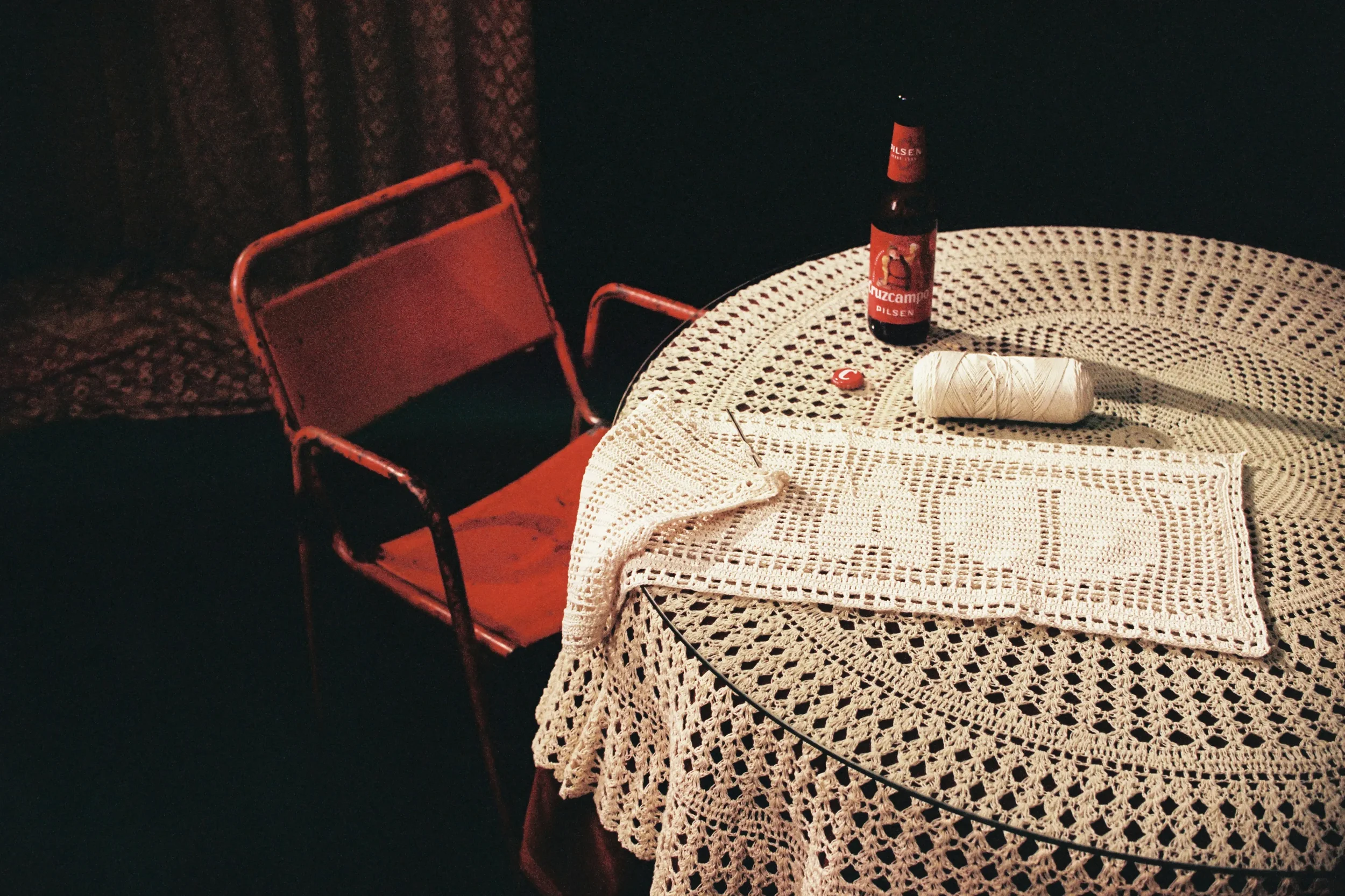 Mesa redonda con mantel de crochet blanco, sobre la cual hay una botella de cerveza con etiqueta roja, un ovillo de hilo blanco, una aguja y un botón rojo, y una silla metálica de color naranja a la izquierda. Al fondo, cortinas oscuras con patrón.
