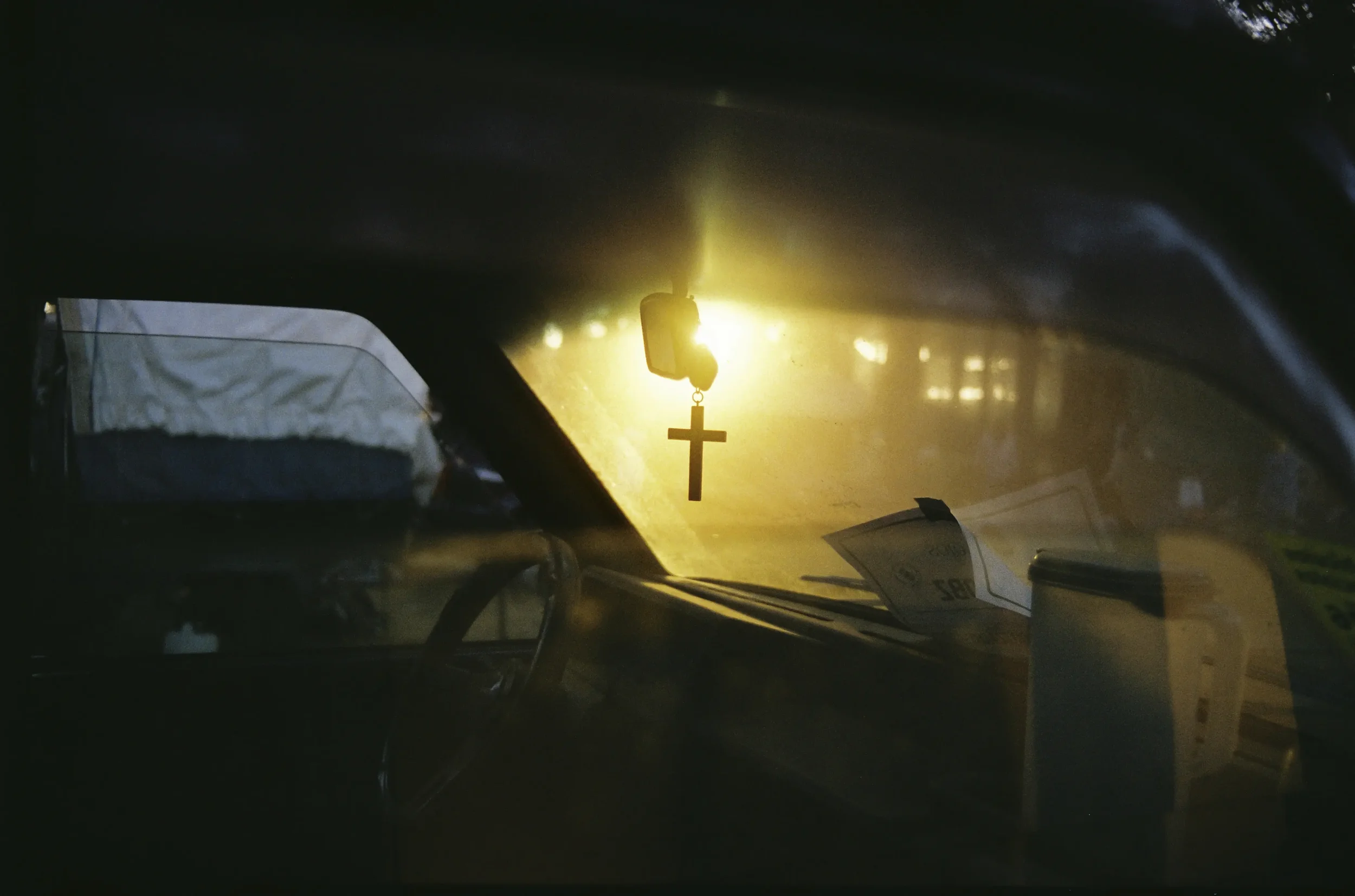 Interior de un vehículo con un crucifijo colgado del espejo retrovisor y luz brillante desde el parabrisas trasero, con objetos y papeles en el tablero y el asiento de pasajero.