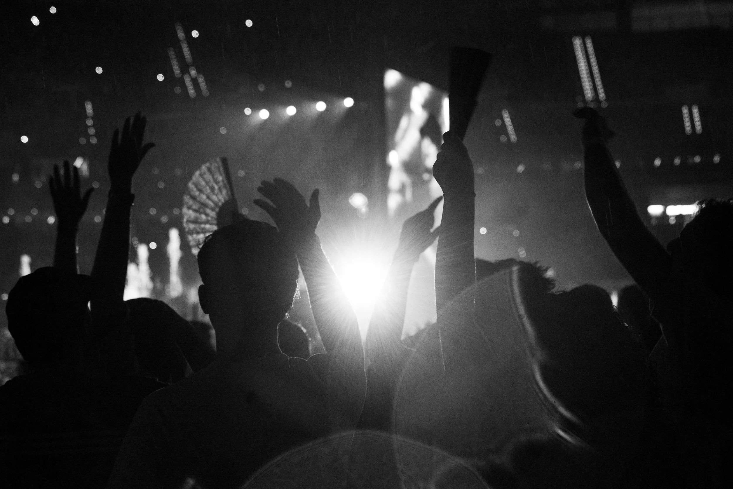 Personas levantando las manos en un concierto en un lugar oscuro con luces y una pantalla grande de fondo.