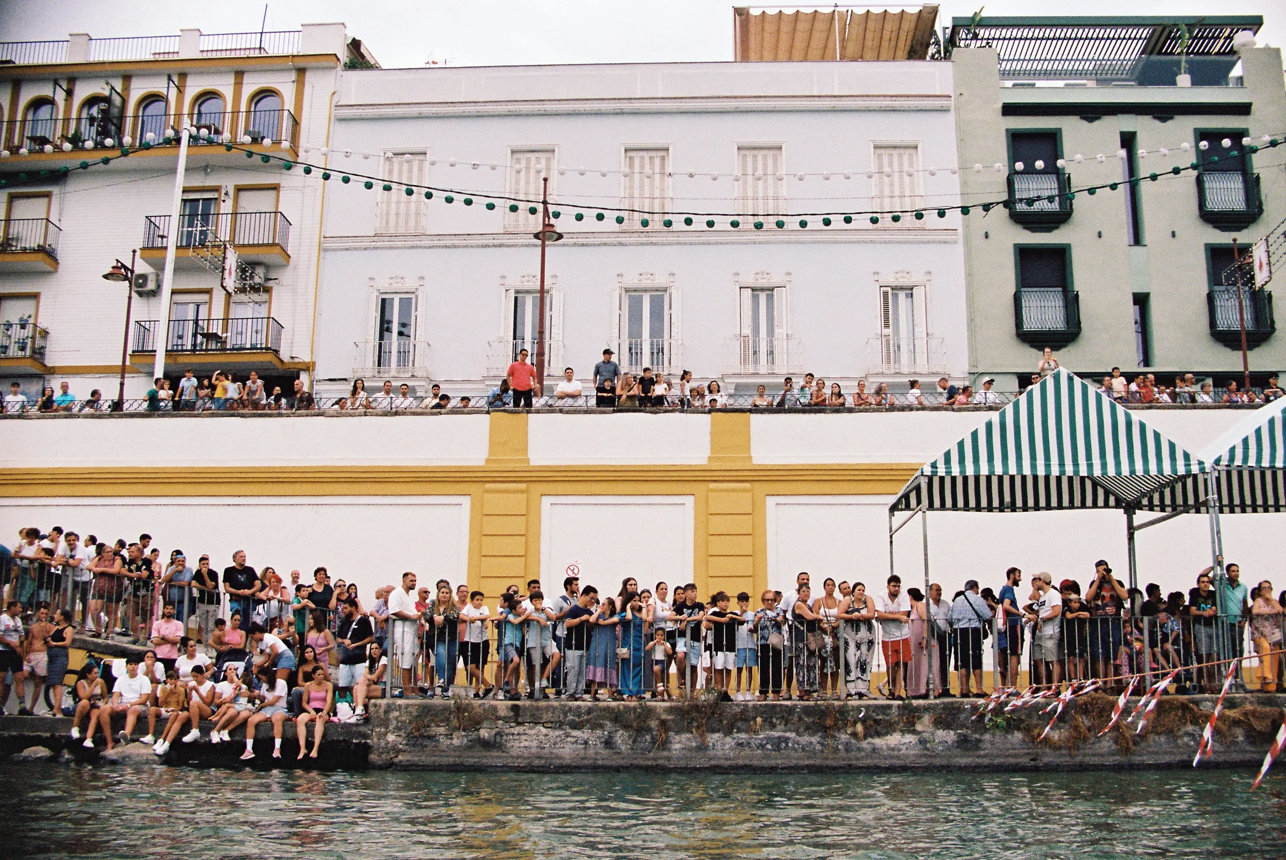 Personas esperando en una fila junto a un muelle más allá de la cual hay edifiicios con balcones y luces decorativas colgadas, en una escena urbana durante el día.
