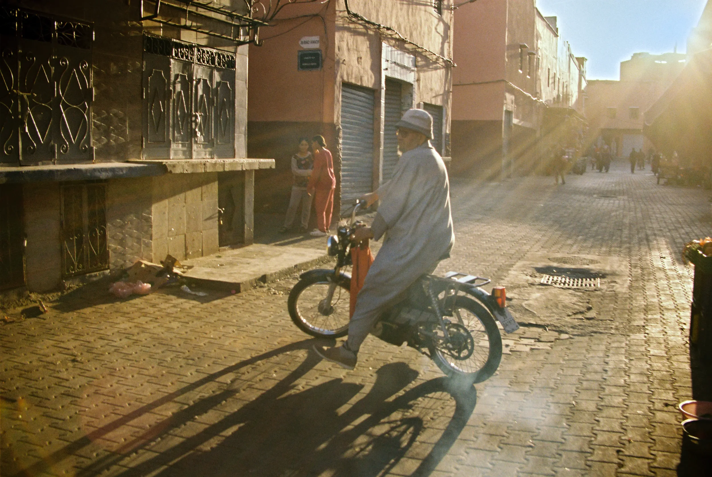 Persona adulta en bicicleta en calle de ciudad, con edificios y otros peatones al fondo, con luz cálida del sol