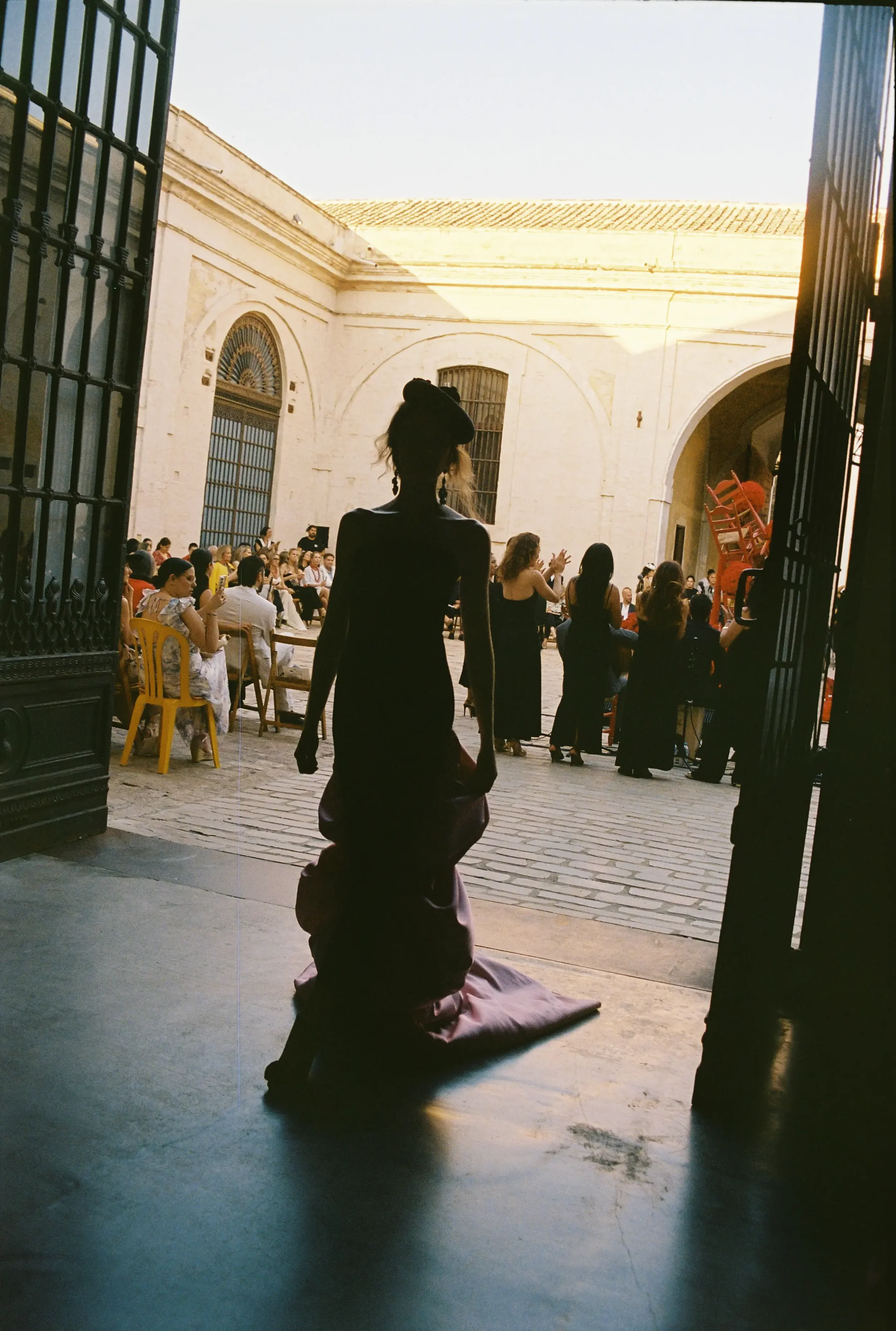 Mujer en vestido largo, plana en tapete, mirando hacia un patio con gente, sillas amarillas y un edificio con ventanas con rejas, al fondo la luz del día.