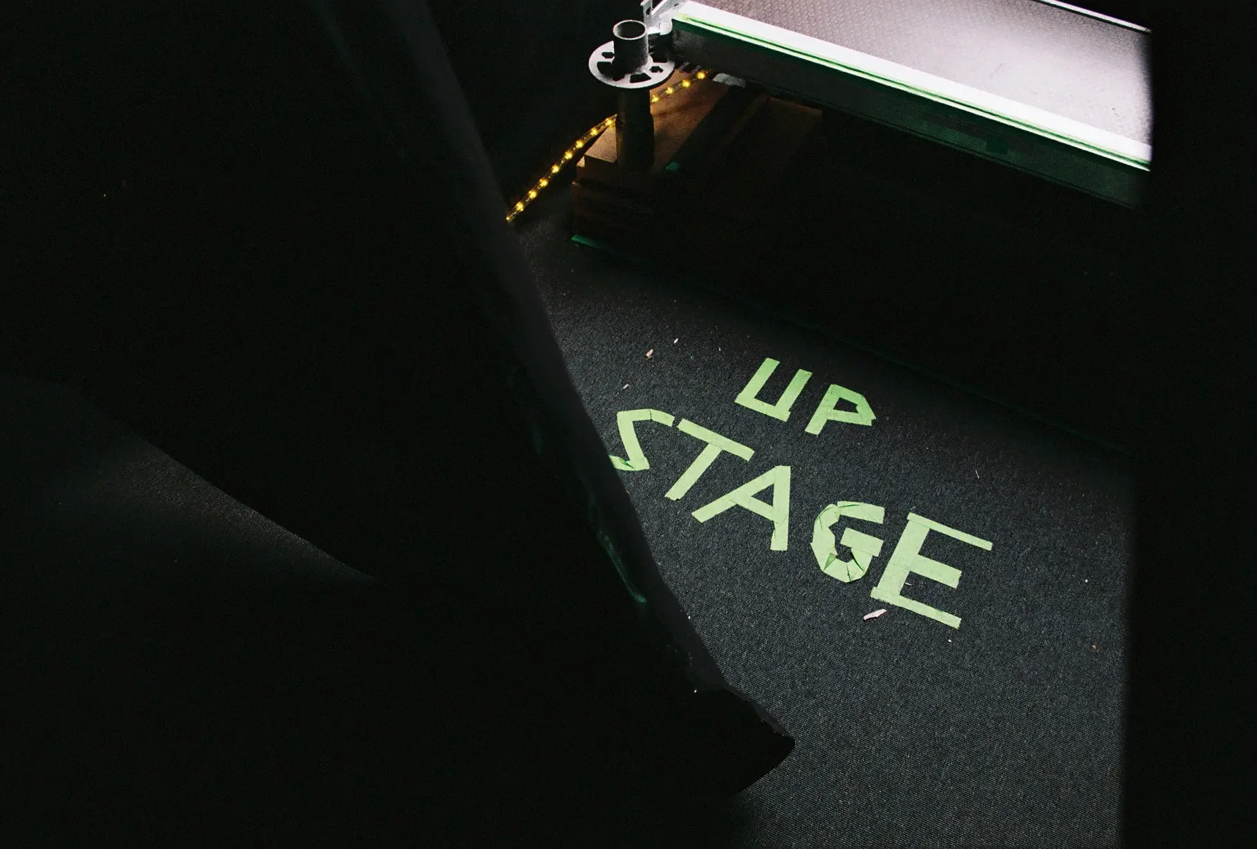 Plano de una oficina con una alfombra que dice 'UP STAGE' en letras verdes, una mesa de trabajo y una silla visible, con poca iluminación.
