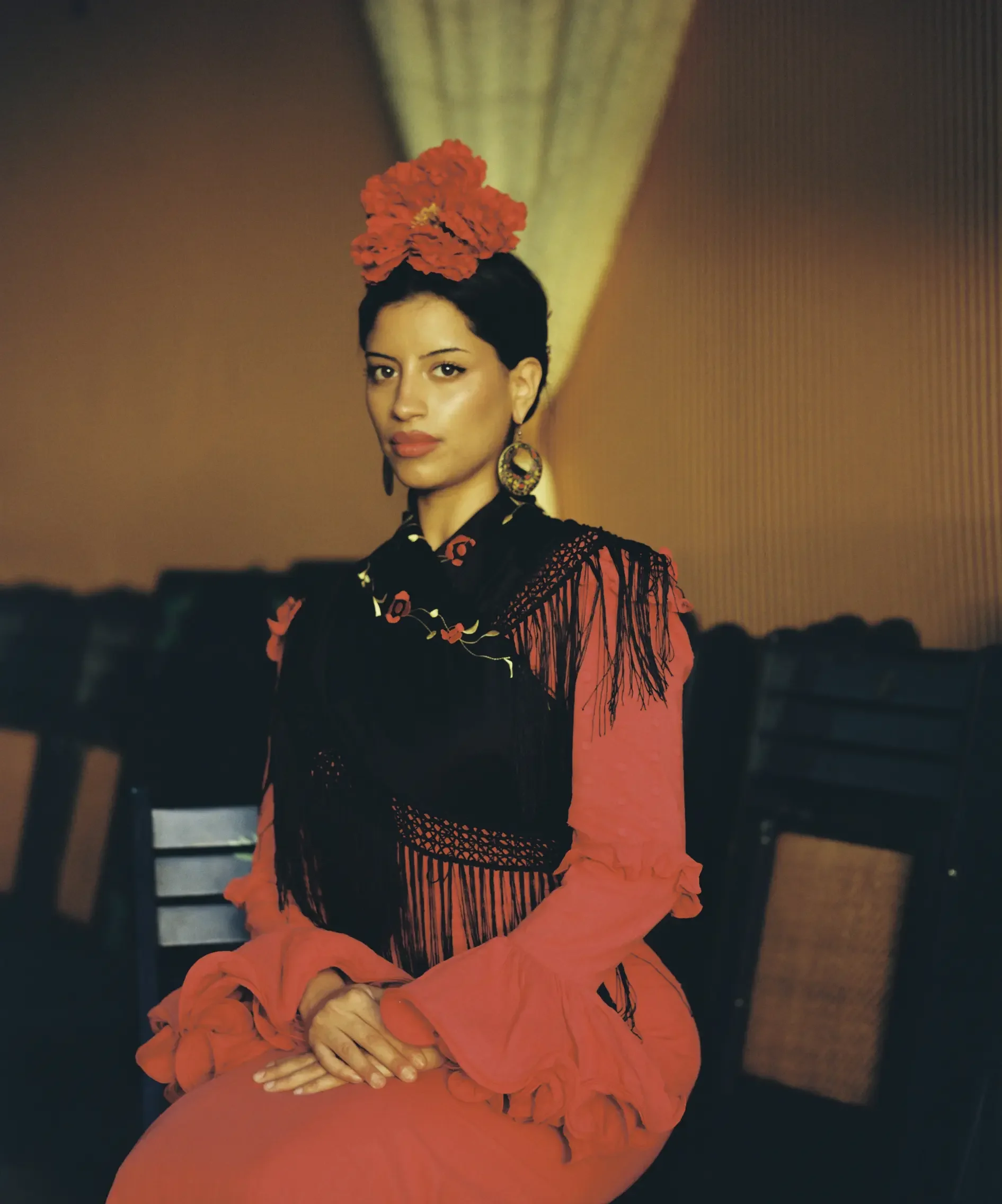 Retrato de una mujer con vestimenta tradicional mexicana, pelo corto y oscuro, luciendo un peinado decorado con una flor grande de color rojo. Ella lleva grandes aretes y un vestido con detalles de encaje y bordados en tonos rojo y negro.