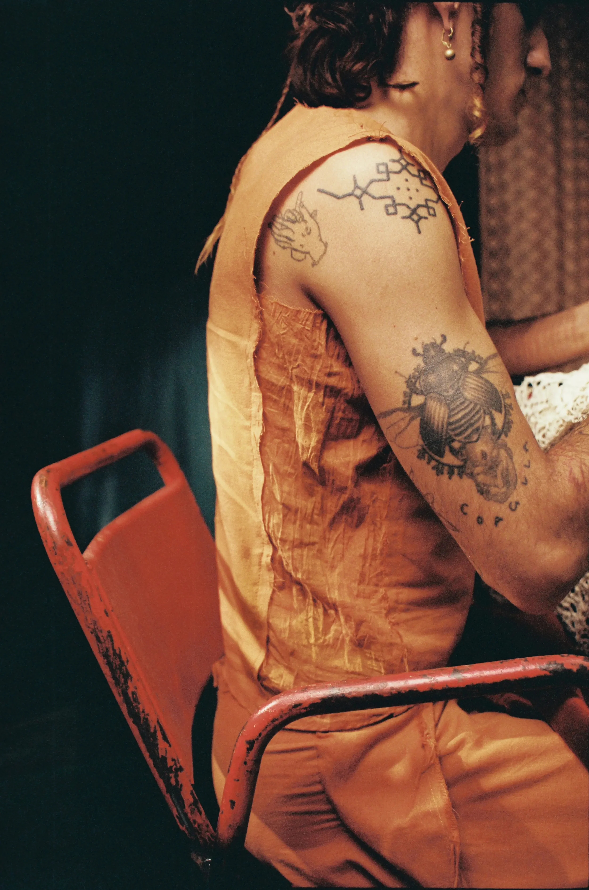 Persona con tatuajes en el brazo, sentada en una silla de metal roja, lleva una camiseta de tirantes de color beige y pantalones cortos de color marrón, ambiente oscuro.