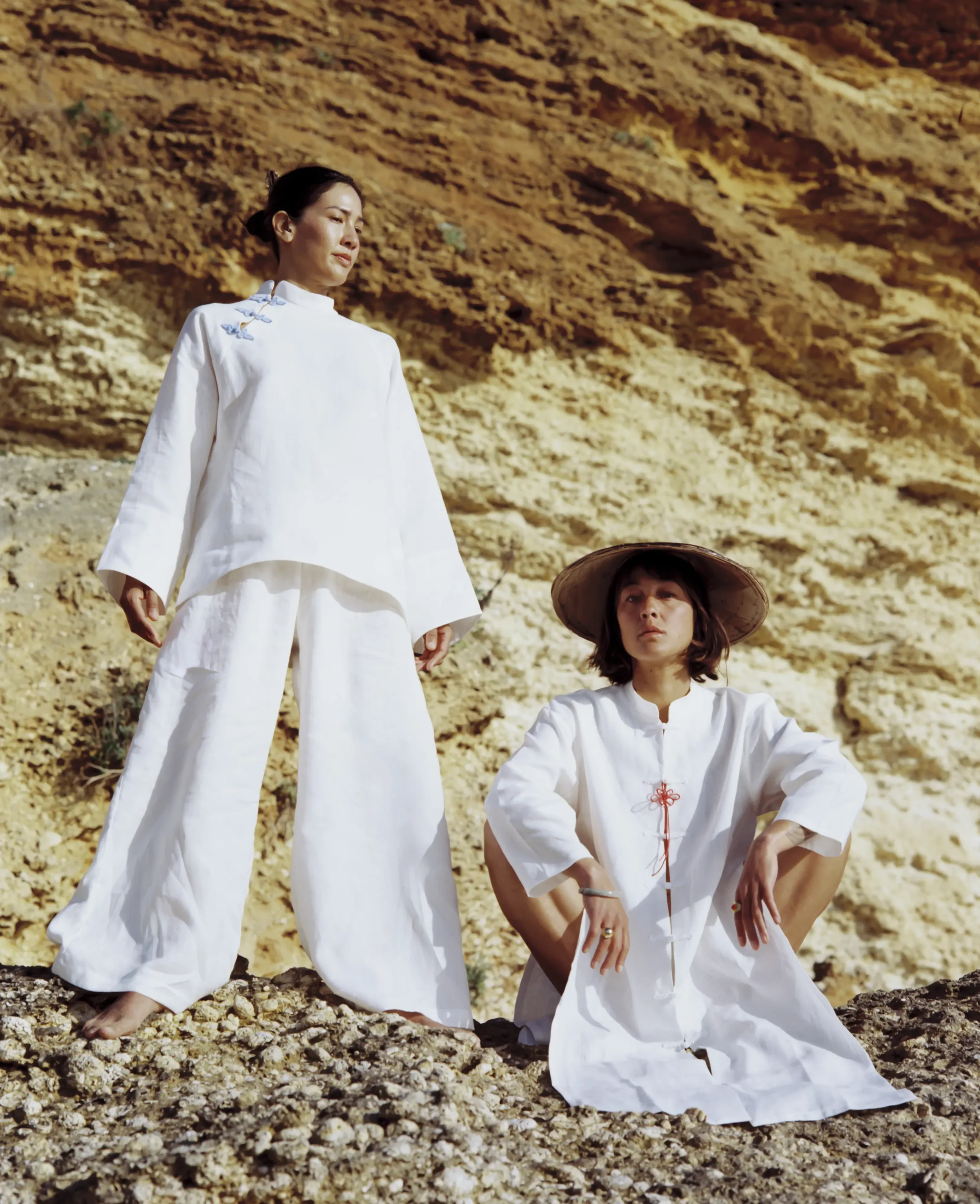 Tres personas con ropa blanca en un paisaje de tierra y rocas, mirando pensativas y sosteniendo posturas meditativas.