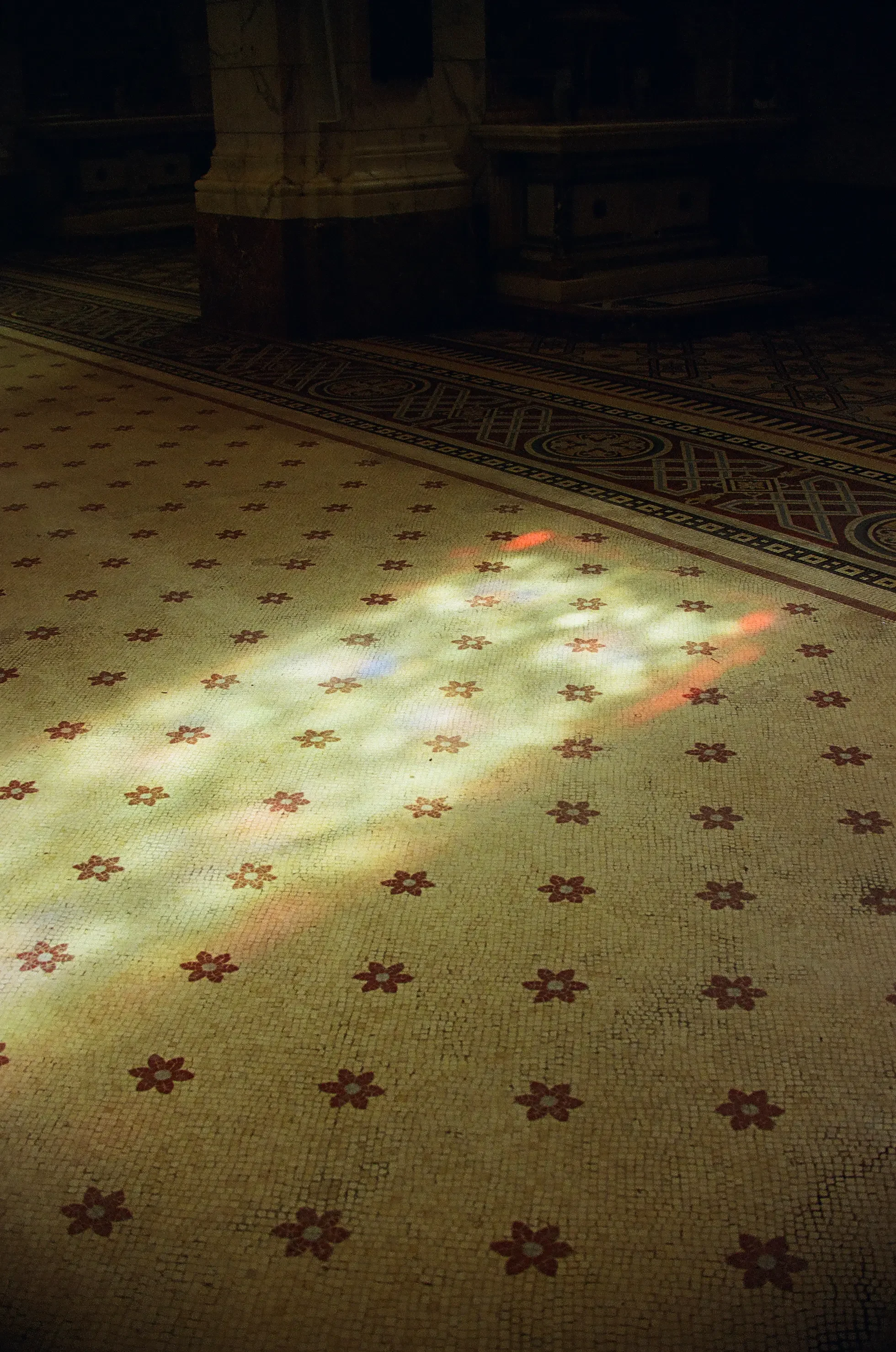 Suelo con mosaico de flores rojas y fondo beige, y parte de un piso con un diseño geométrico y bordes en tonos oscuros, en un espacio con poca iluminación.