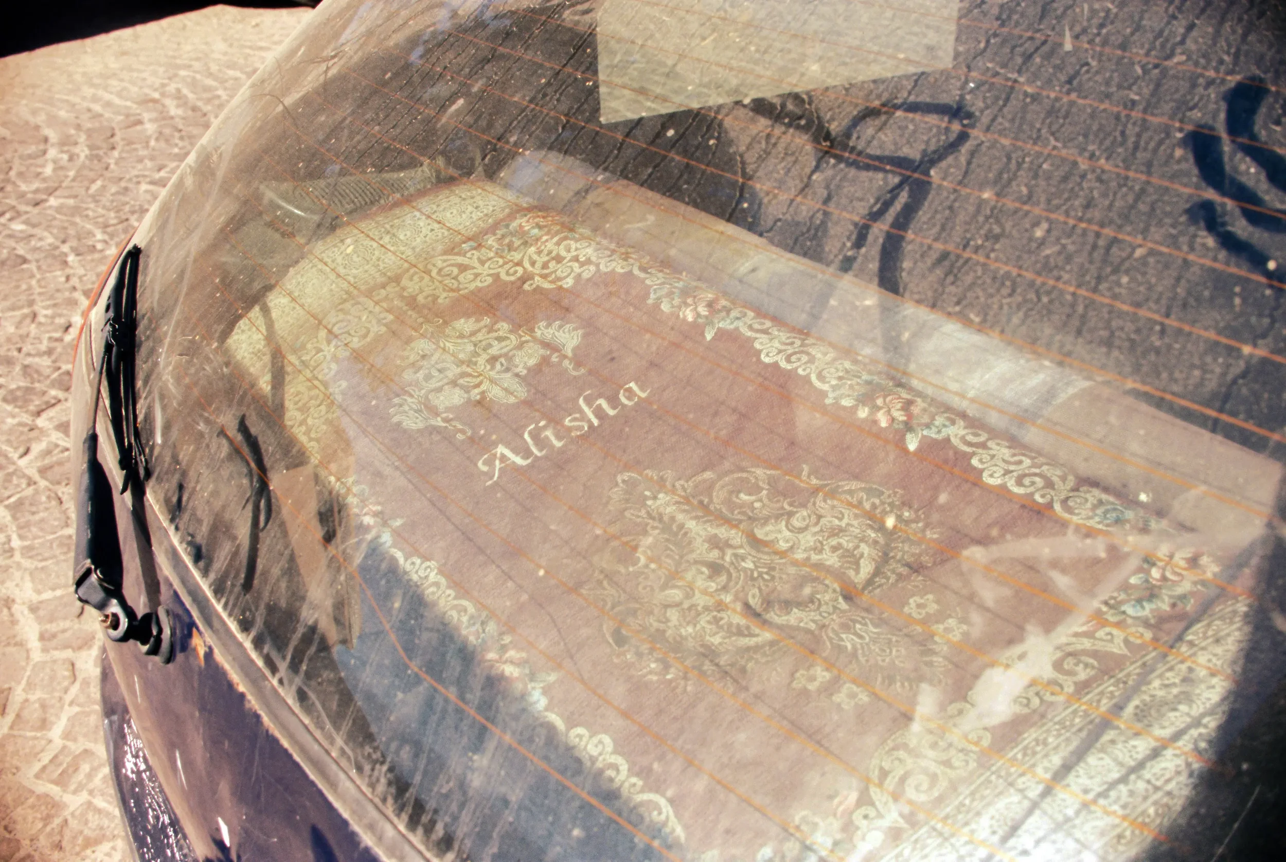 El interior de un automóvil con una caja de madera decorada y la palabra 'Alisha' grabada, visible a través del vidrio trasero, en un área exterior con suelo de piedra.