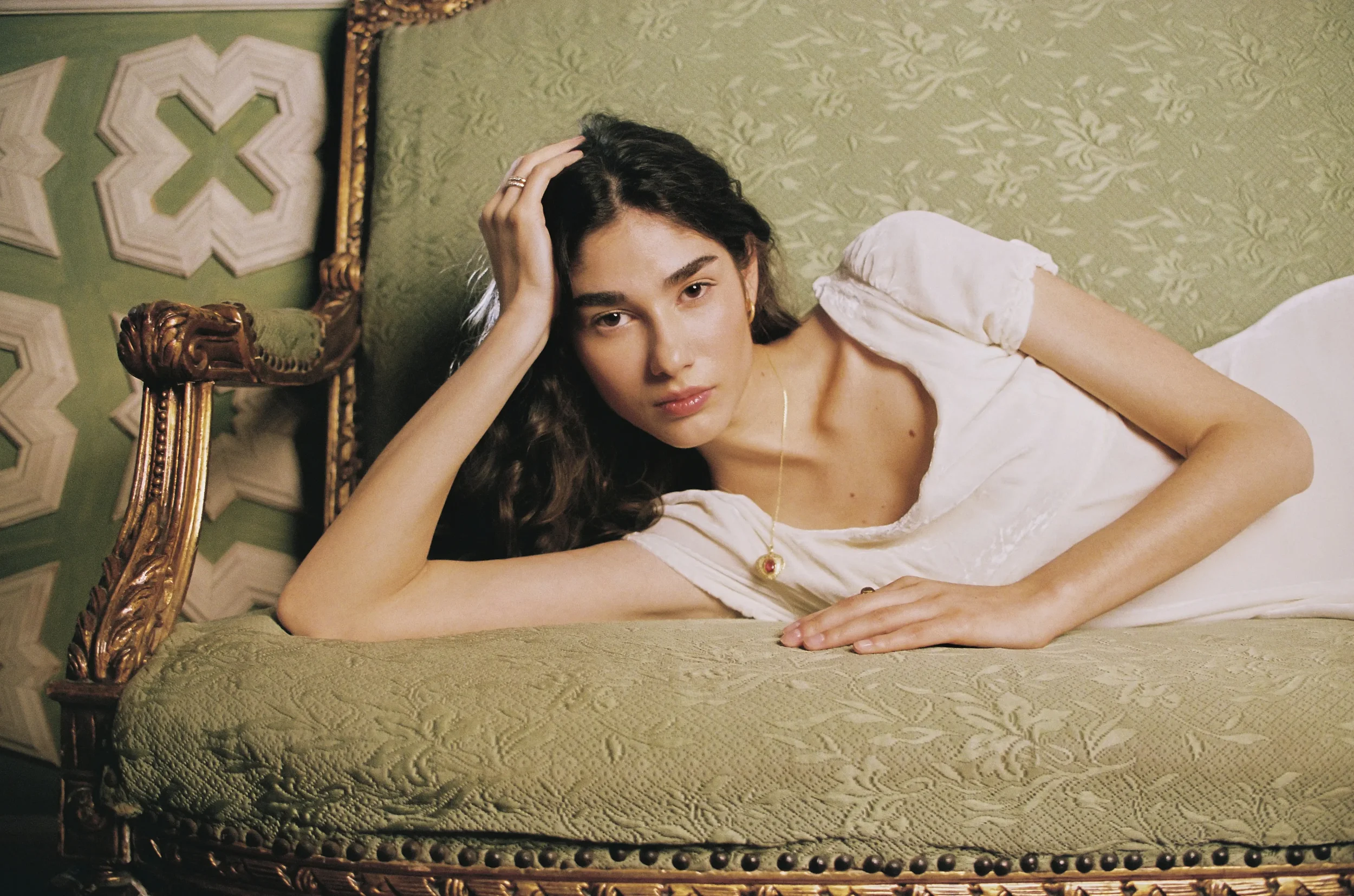 Joven mujer recostada en una cama antigua, con cabecera de madera tallada y tapizada en tela verde con un patrón floral, vestida con un vestido blanco y usando un collar con un pendant redondo, tocada con cabello rizado y mirando hacia la cámara.