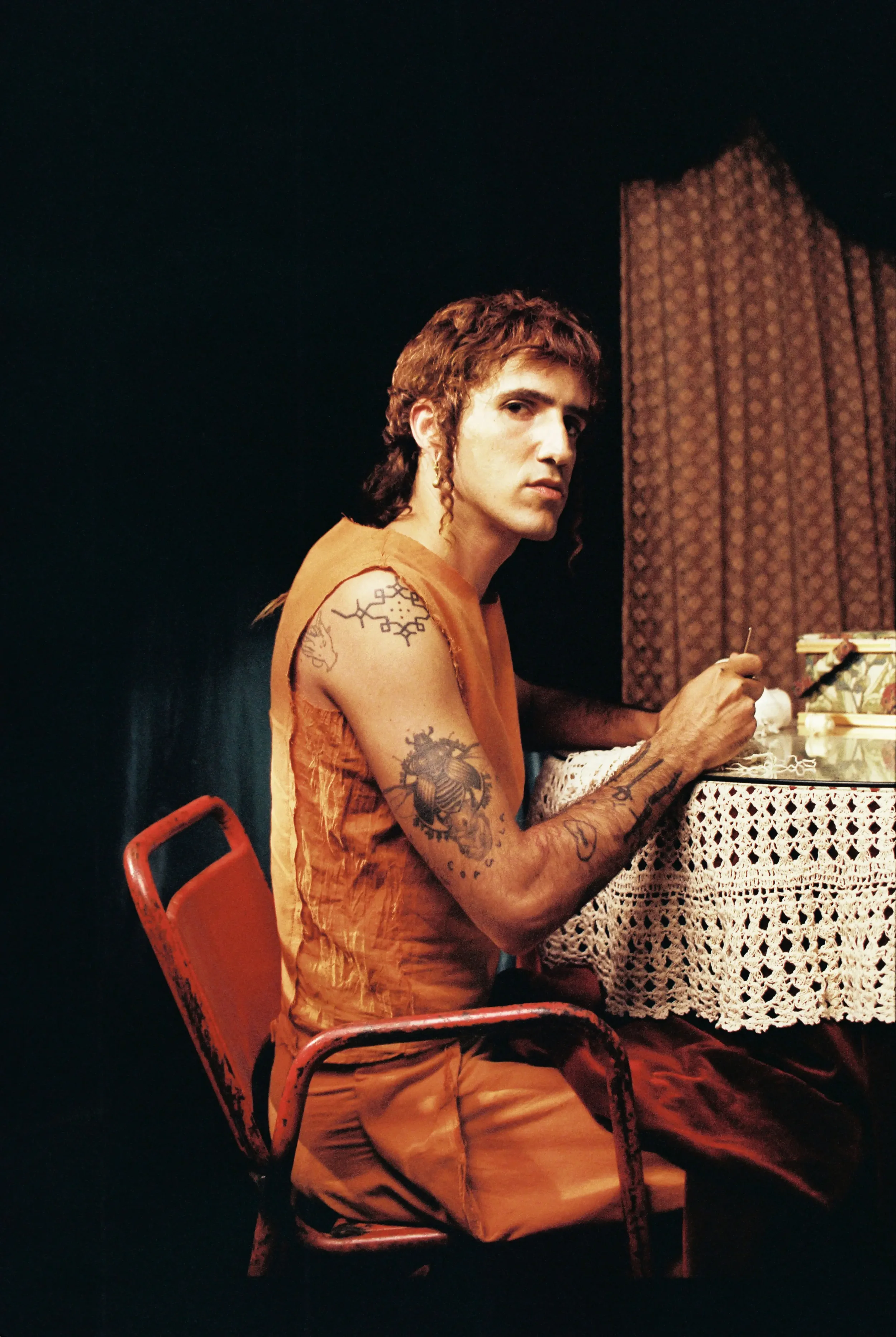 Hombre de cabello rizado y tatuajes sentado en una silla roja, sosteniendo un pincel y pintando en una mesa con mantel de encaje, fondo oscuro y cortina marrón.