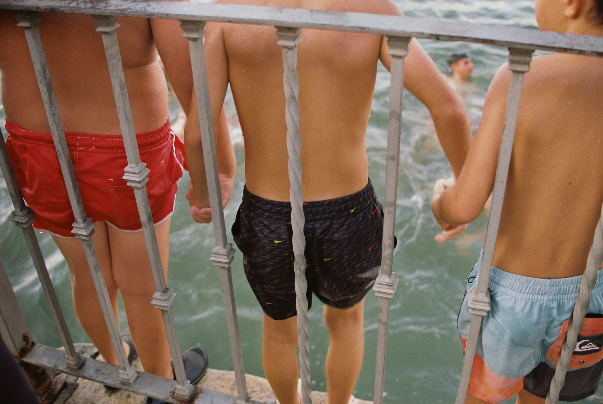 Jóvenes de espaldas, sin camiseta y con ropa deportiva, en una especie de muro de seguridad frente al mar, algunos con gotas de agua en la piel, observando el agua.
