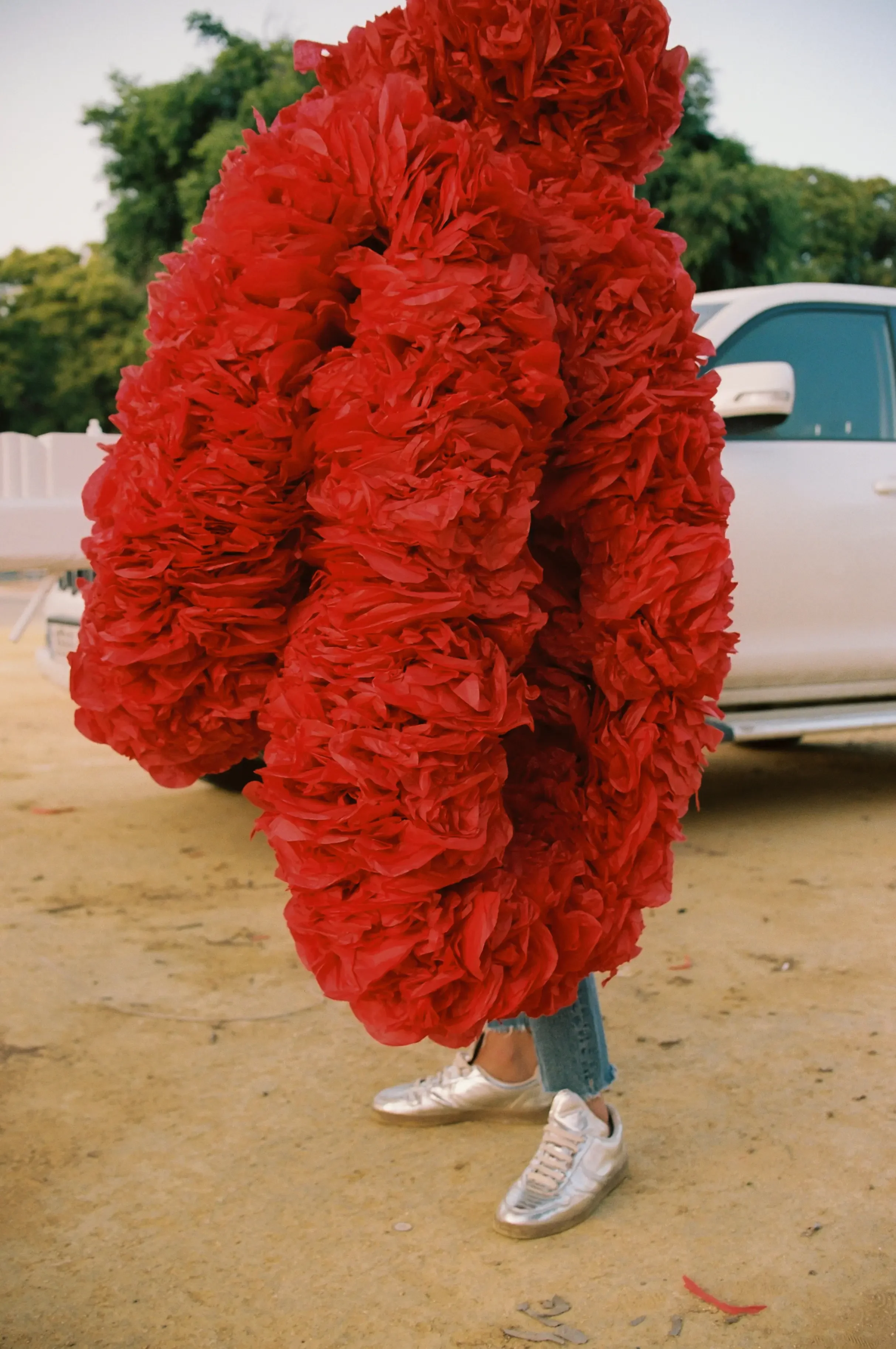 Persona con pantalones cortos y tenis blancos, sosteniendo un disfraz de flores rojas en forma de corazón, en un entorno al aire libre con árboles y un vehículo blanco en el fondo.