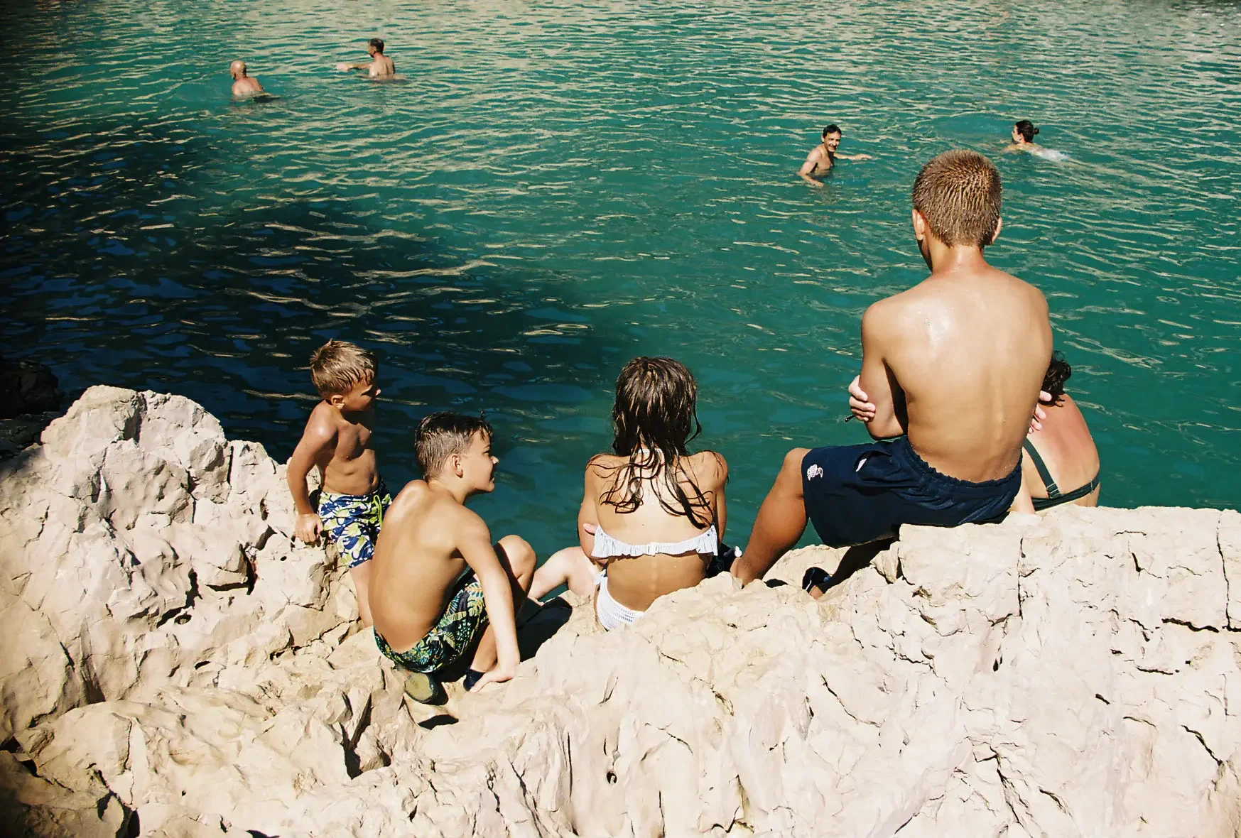 Grupo de niños y adultos sentados en una roca junto al agua, algunos en el agua y otros observando, en un día soleado en la playa.