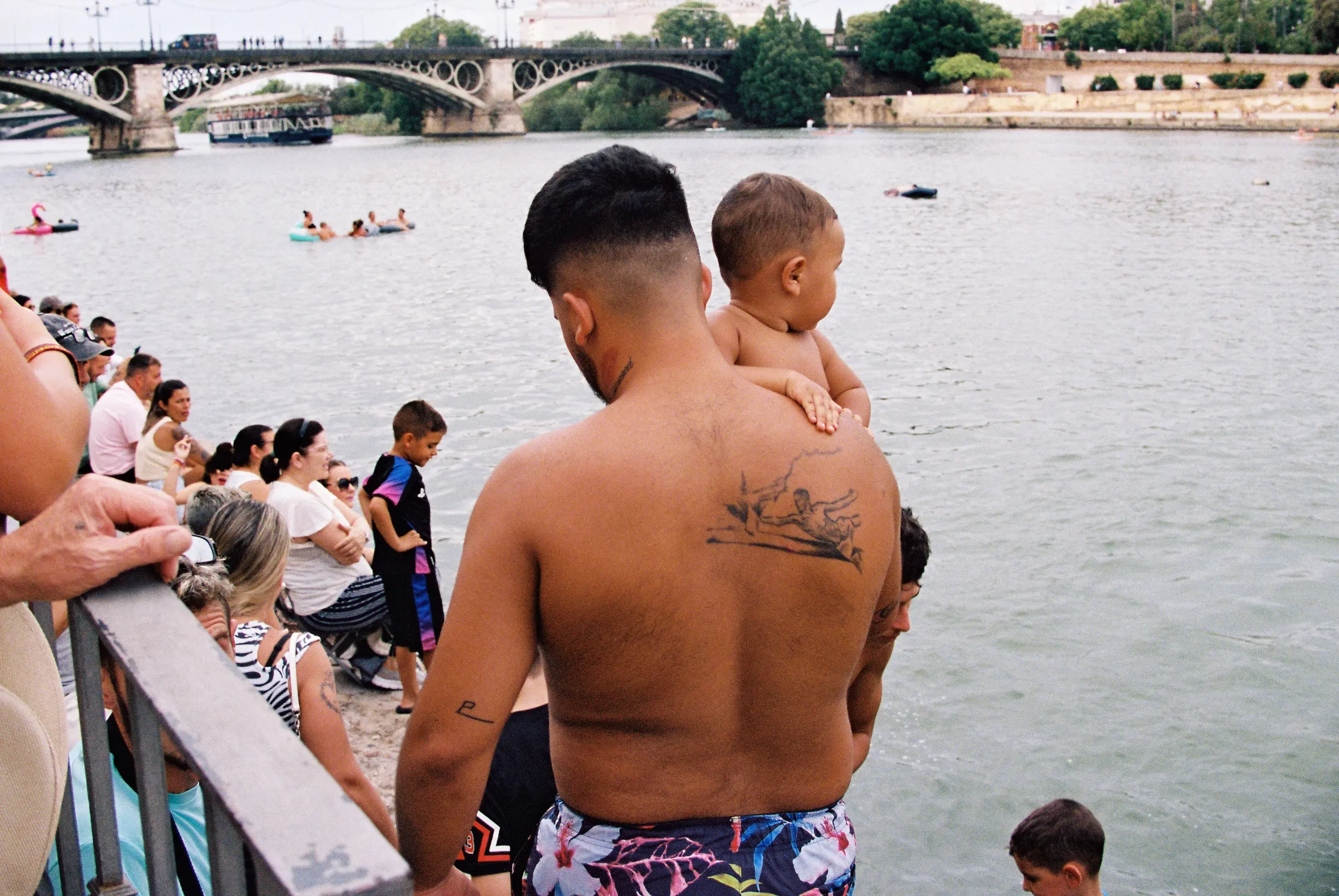 Un hombre sin camiseta con tatuajes sostiene a un niño en brazos mientras observa el río, rodeado de otras personas en la orilla, con un puente y árboles de fondo.